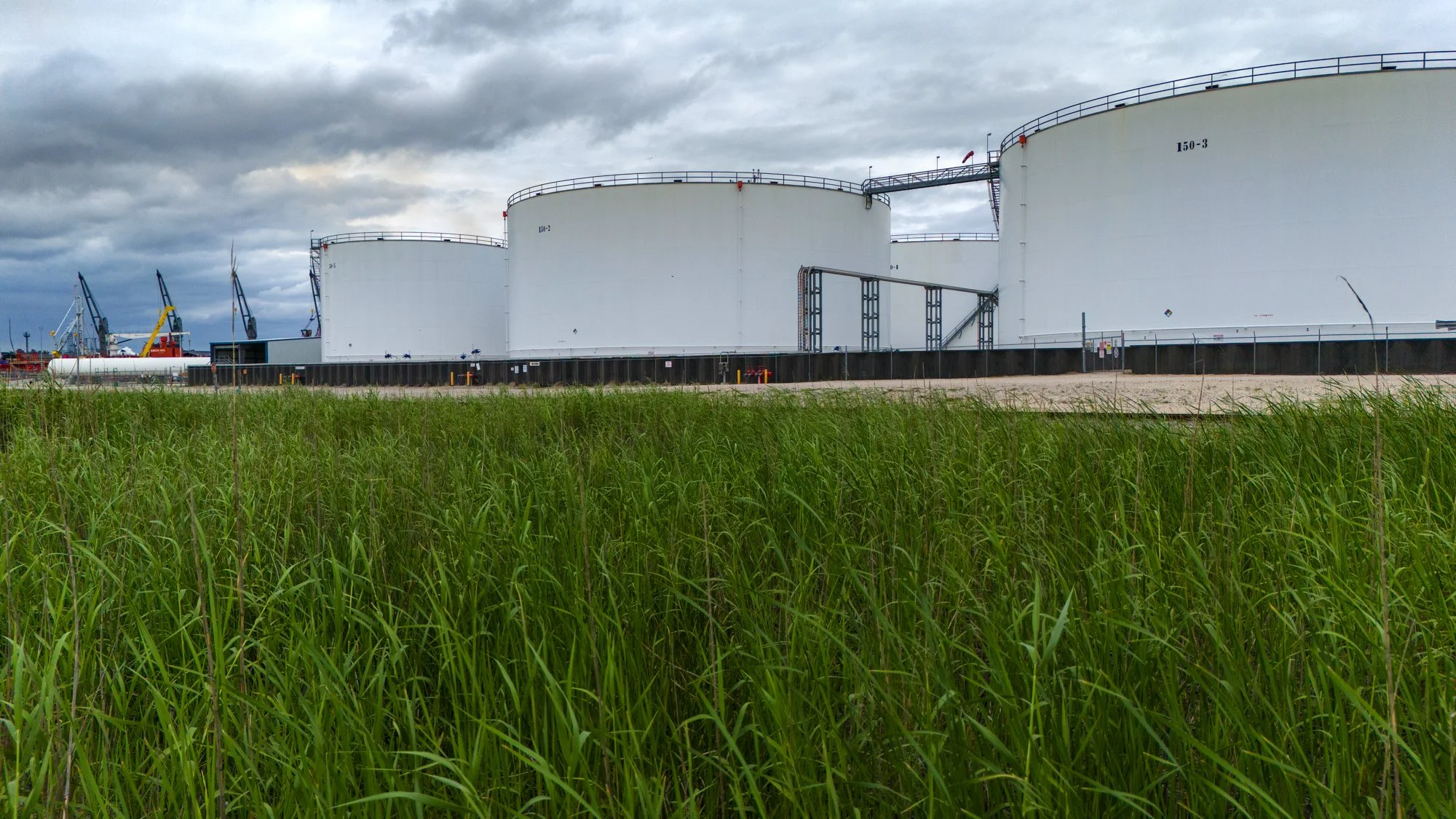 Large white oil storage tanks on a concrete platform behind a field of green grass with cloudy sky in the background.