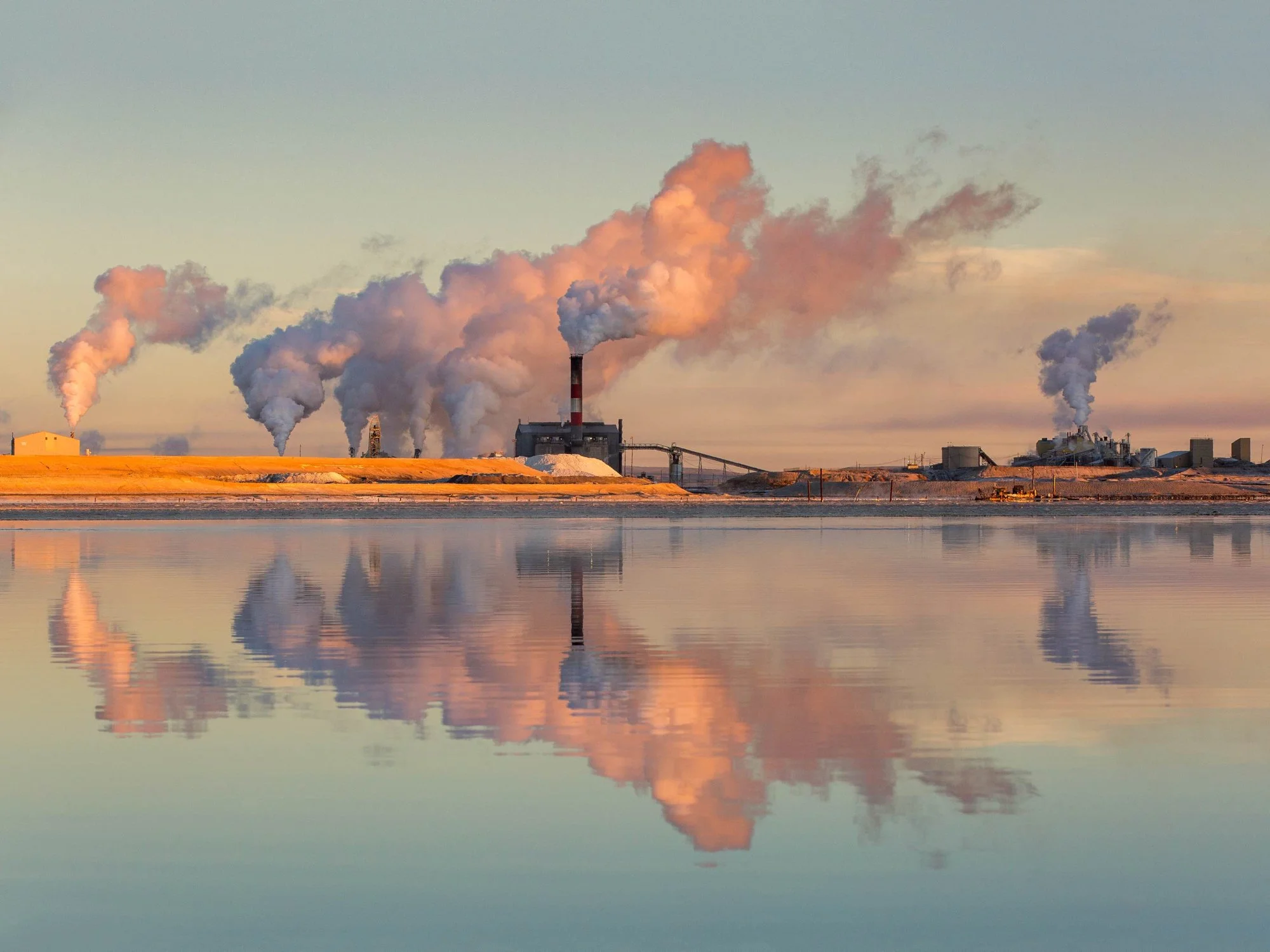 Industrial factory emitting smoke near a calm body of water, with its reflection visible on the water's surface.