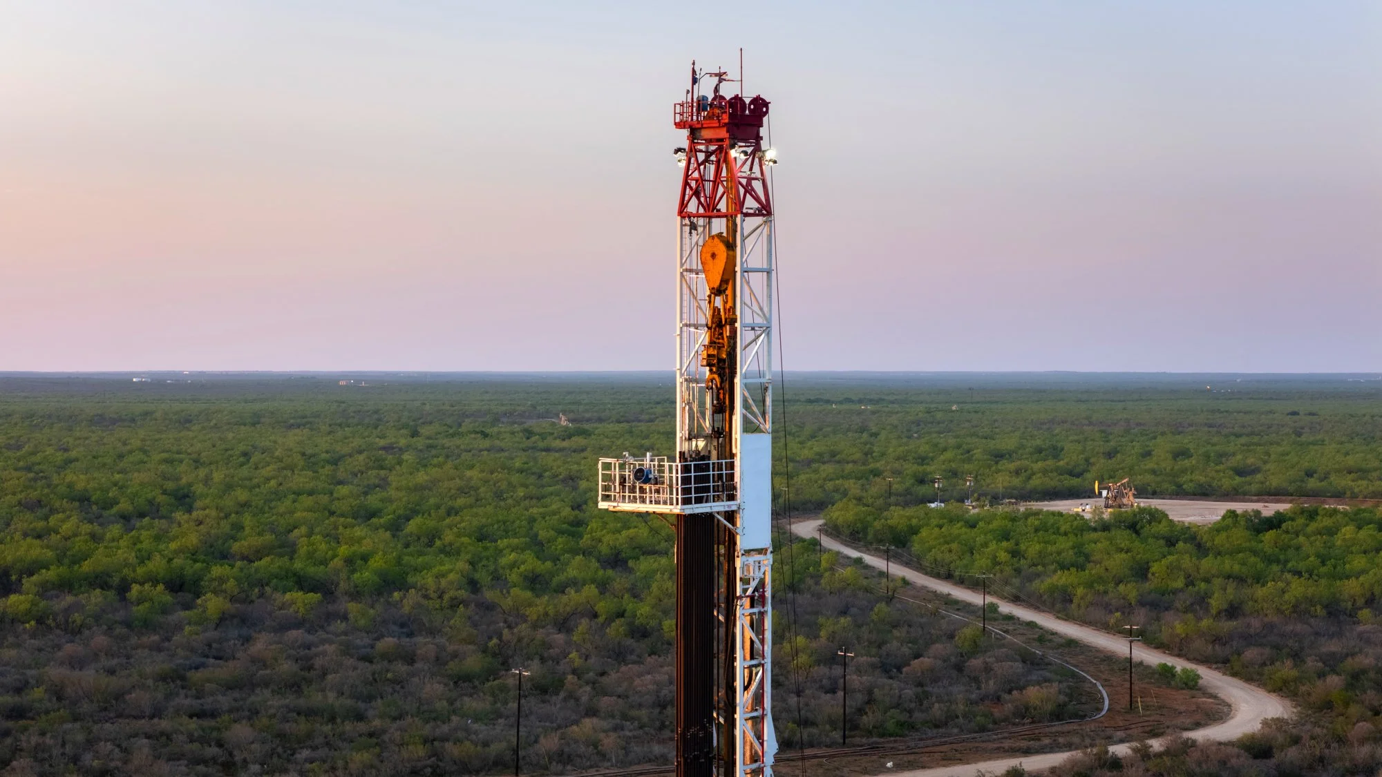 Tall red and white oil drilling rig in a flat, green, rural landscape with dirt roads and sparse trees