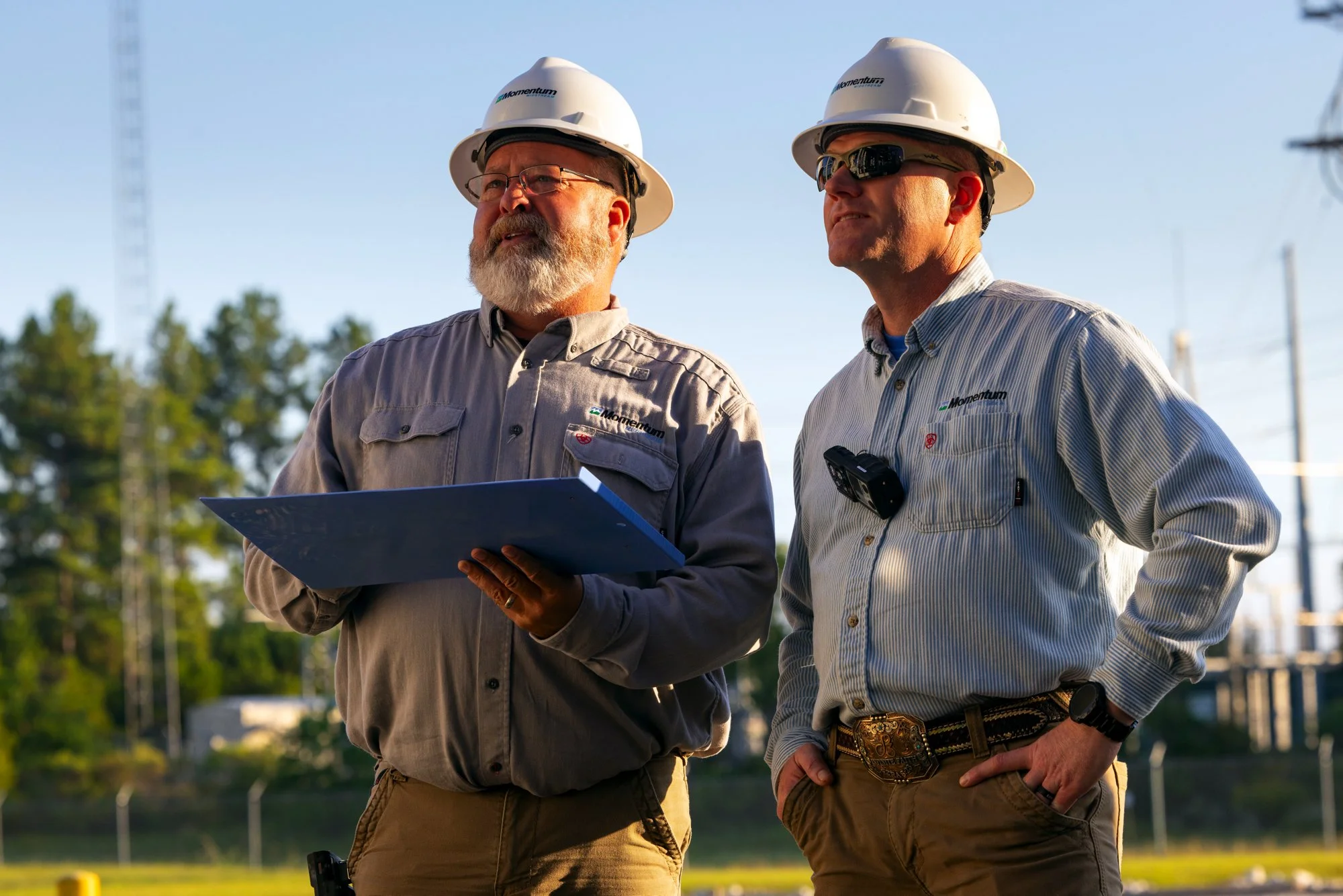 Two male engineers wearing safety helmets and work shirts, standing outdoors and discussing work plans. One holds a clipboard and the other has a walkie-talkie clipped to his shirt.