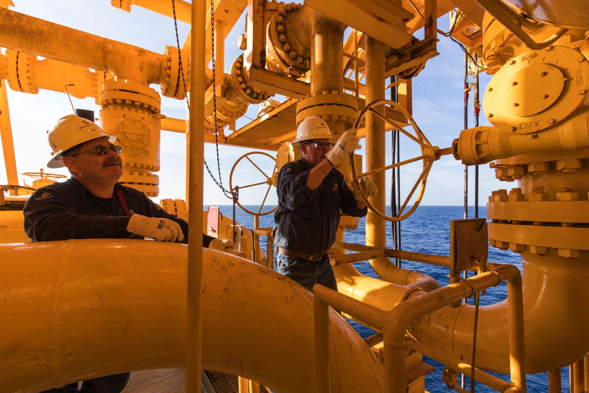 Two workers on an offshore oil platform, wearing helmets and safety gear, working among large yellow pipes with the ocean visible in the background.