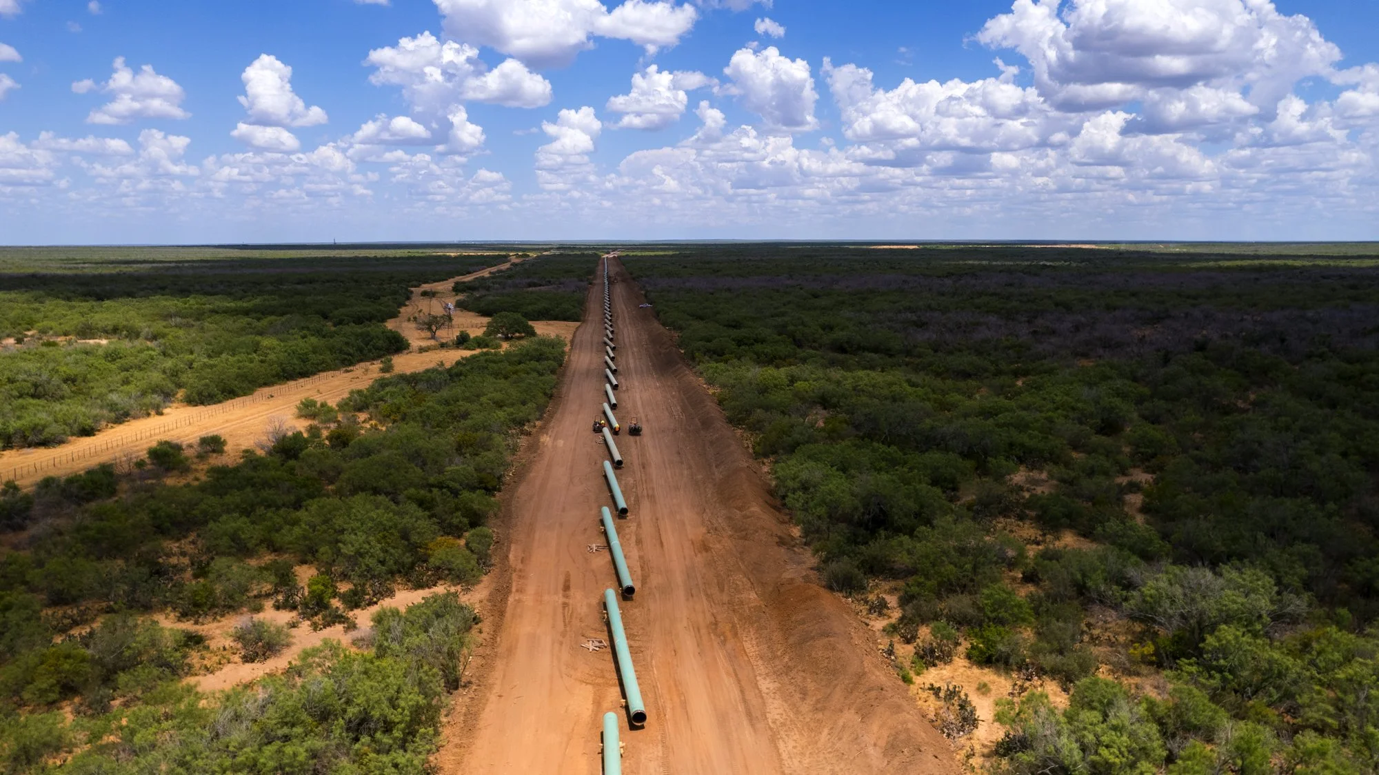 Aerial view of a pipeline construction in a rural landscape with dirt road and green vegetation under a cloudy sky.