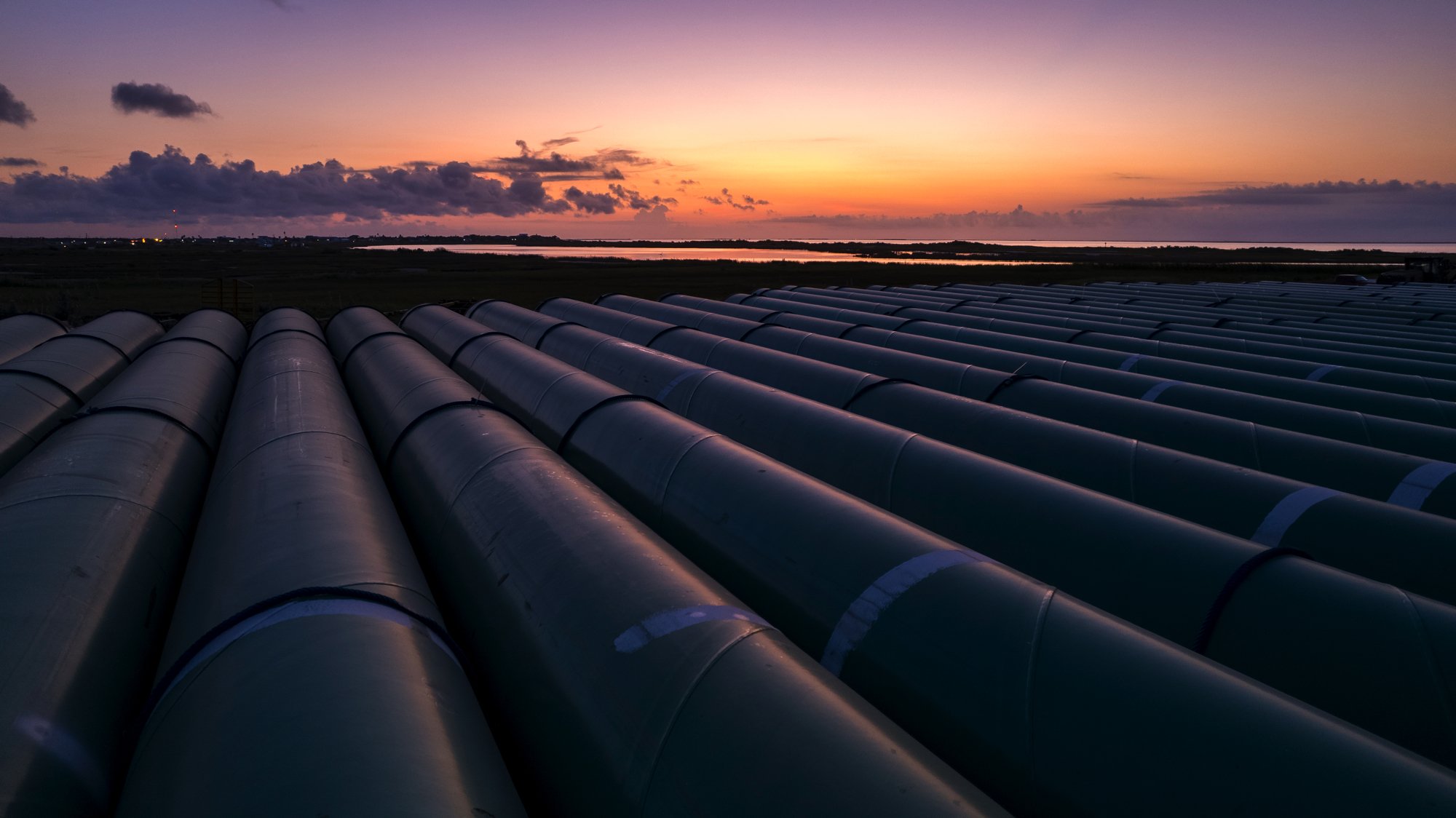 Multiple large industrial pipelines on a field at sunset with water and horizon in the background.