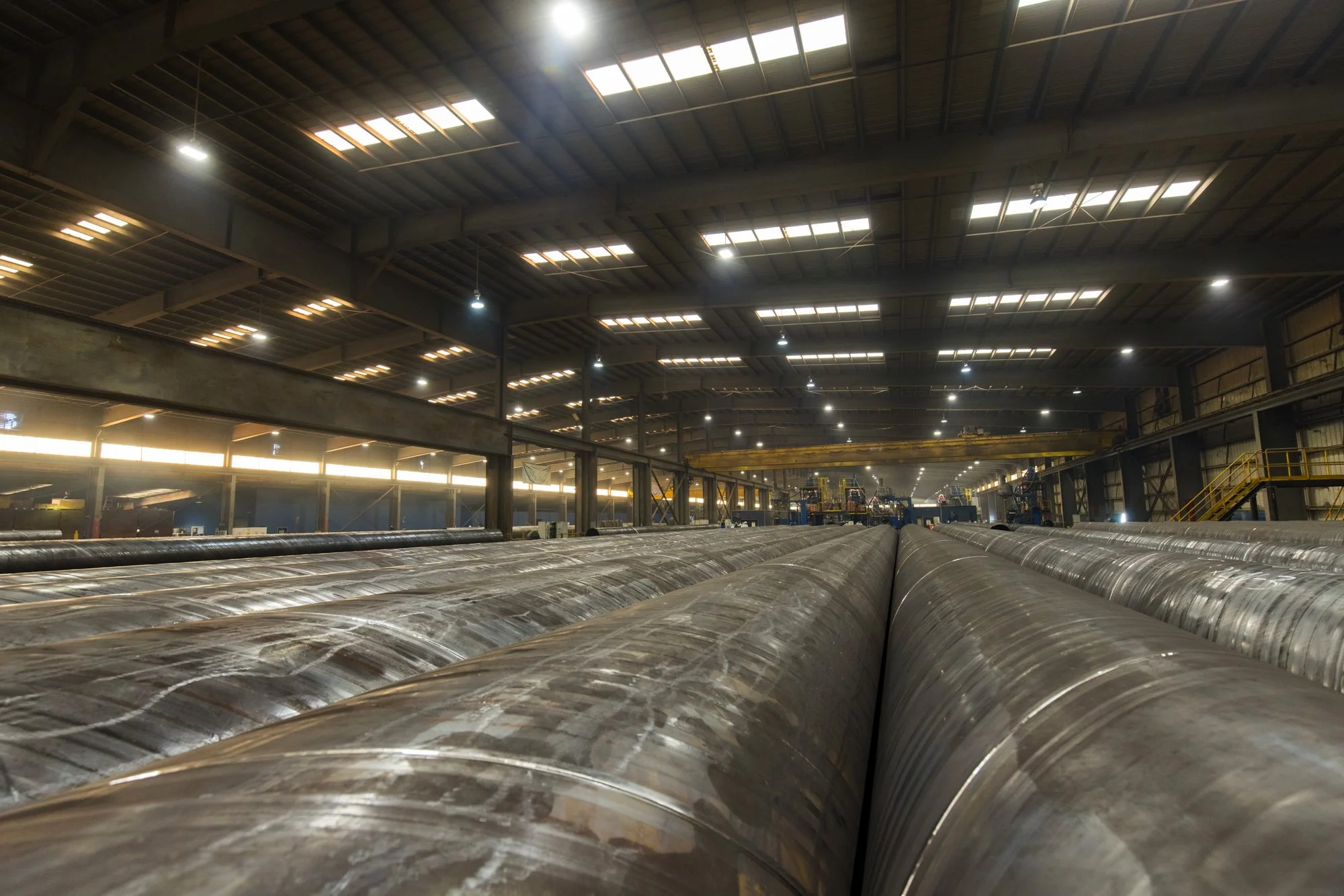 Inside an industrial warehouse with large metal pipes laid out on the floor and overhead cranes in the background.
