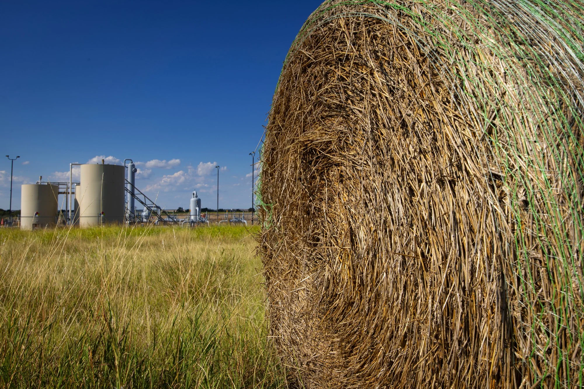 Close-up of a large rolled hay bale in a field with industrial tanks and pipes in the background under a blue sky with scattered clouds.