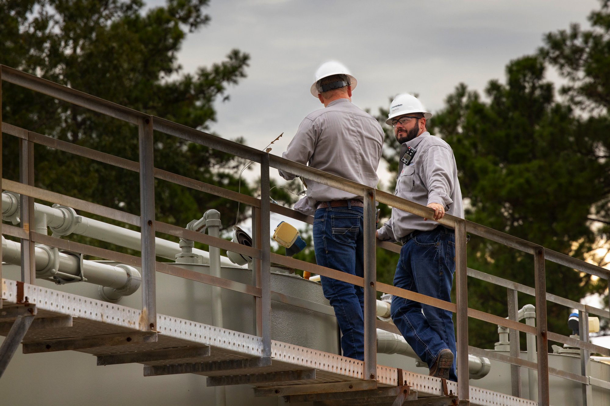 Two industrial workers wearing safety helmets and gray shirts working on a metal platform with pipes, with trees in the background.