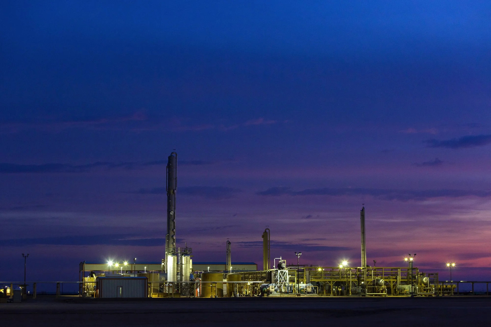 Industrial oil refinery at sunset with purple and blue sky in the background and bright lights illuminating the facility.