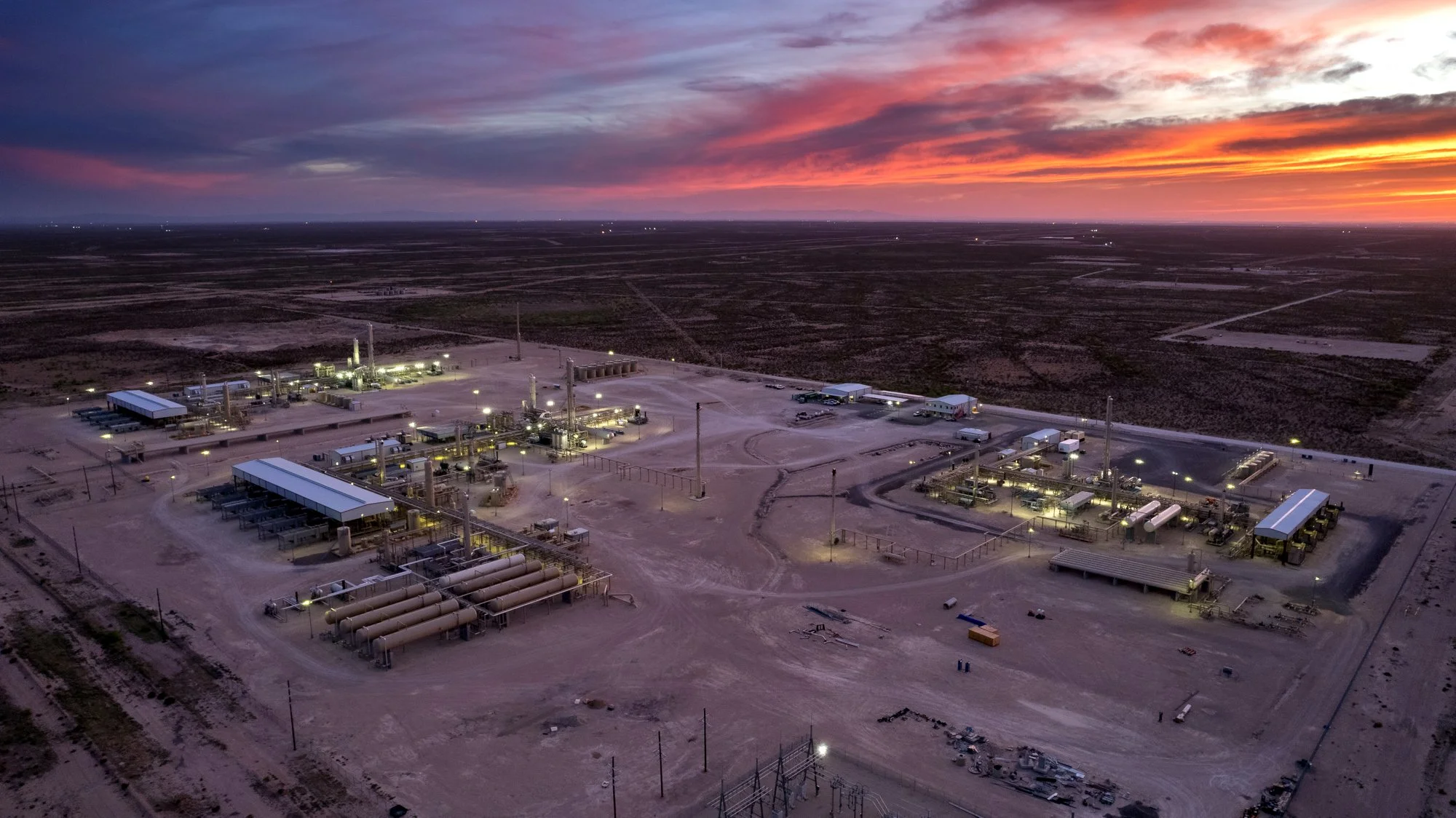A large industrial facility with multiple structures and pipelines illuminated at dusk, surrounded by a flat, desert landscape.