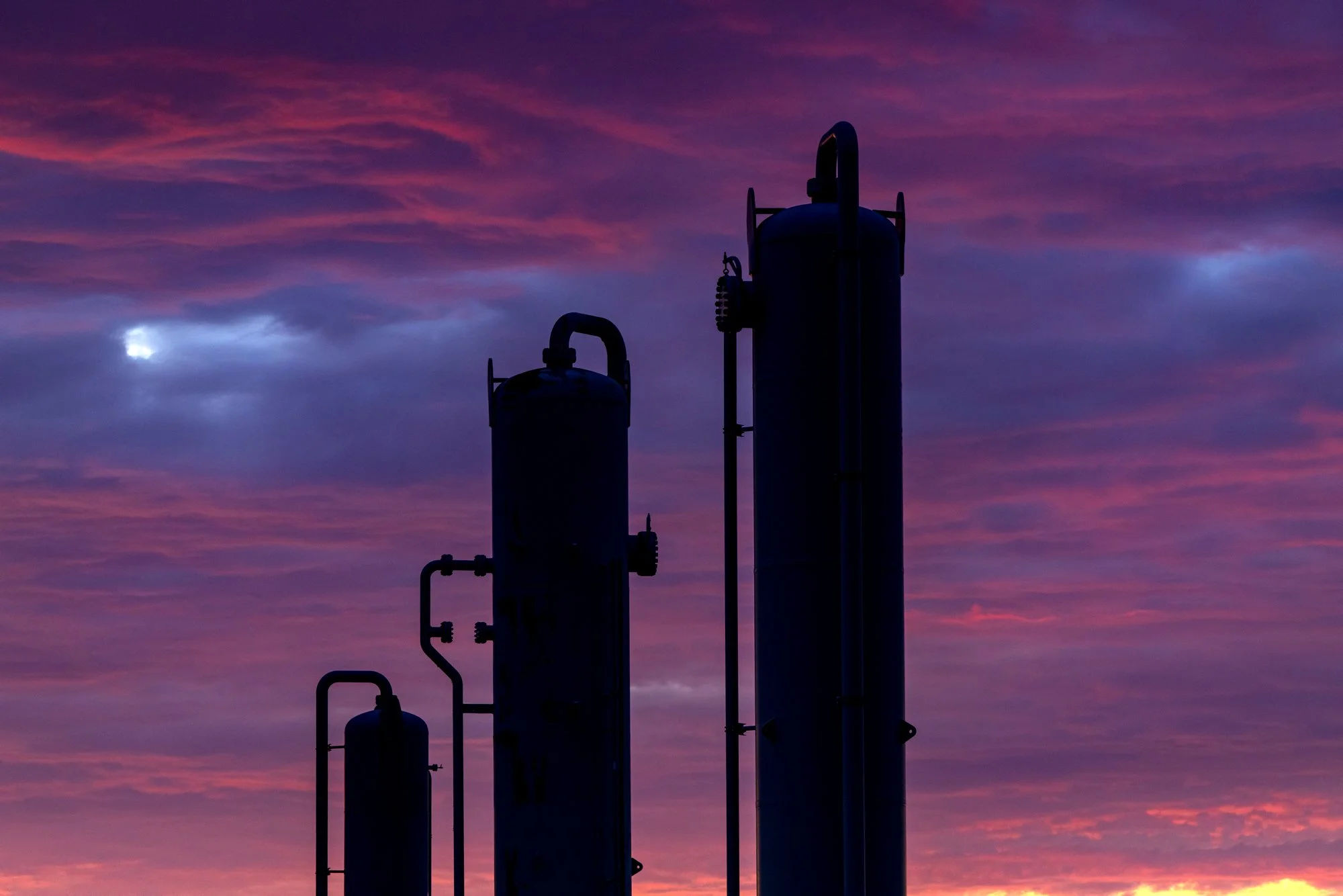 Silhouettes of industrial tanks against a colorful sunset sky with pink and purple clouds.