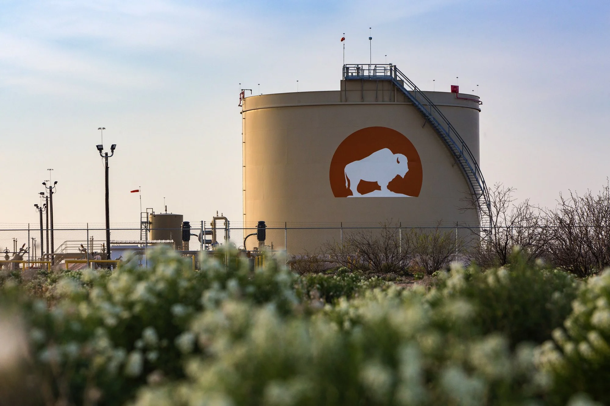 Large industrial storage tank with a white buffalo silhouette logo inside an orange circle. The tank is behind a chain-link fence, with pipes, lights, and trees in the foreground and a clear sky in the background.
