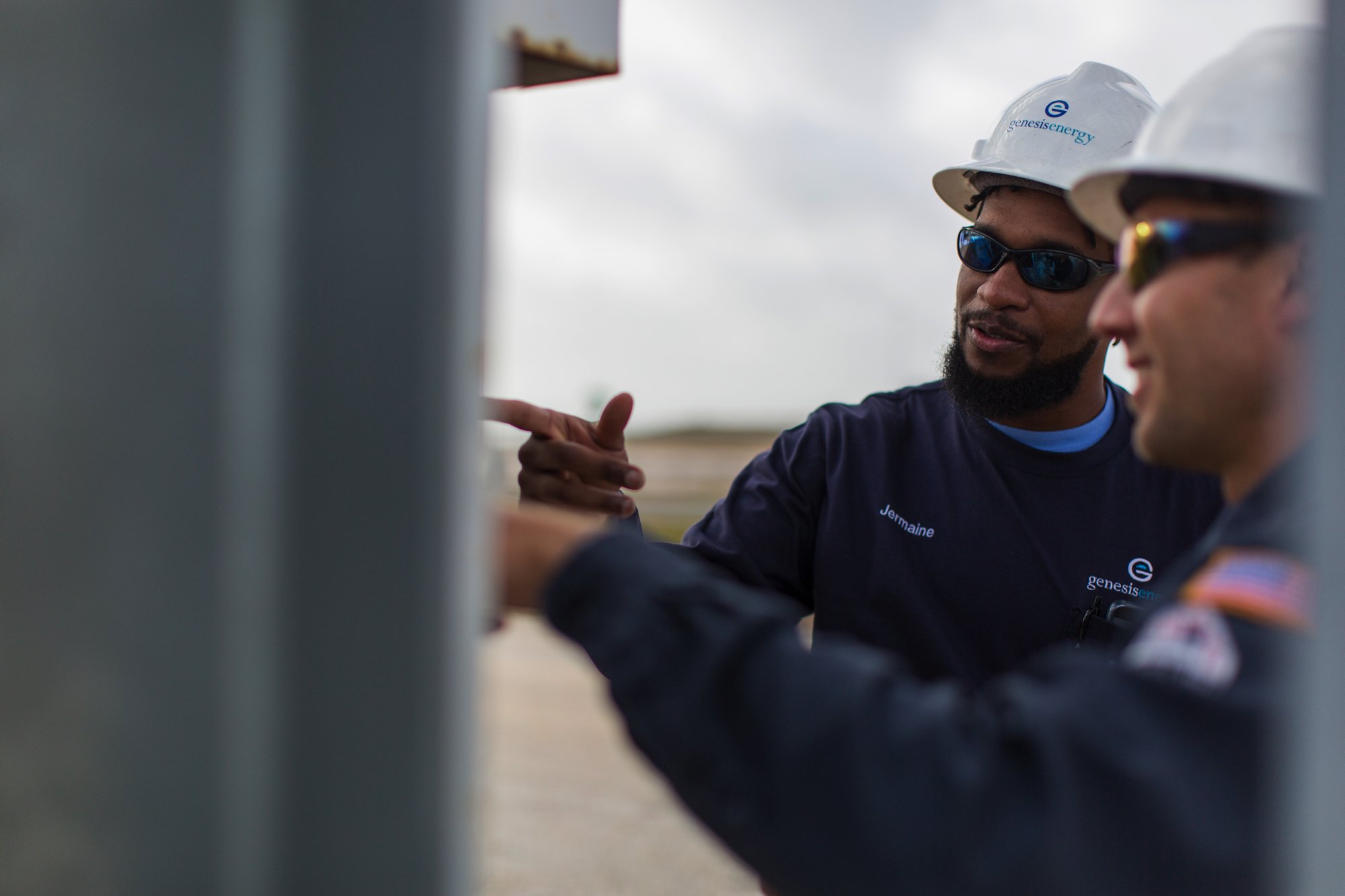 Two engineers wearing Genesis Energy uniforms, safety helmets, and sunglasses discussing work outdoors, with one pointing.