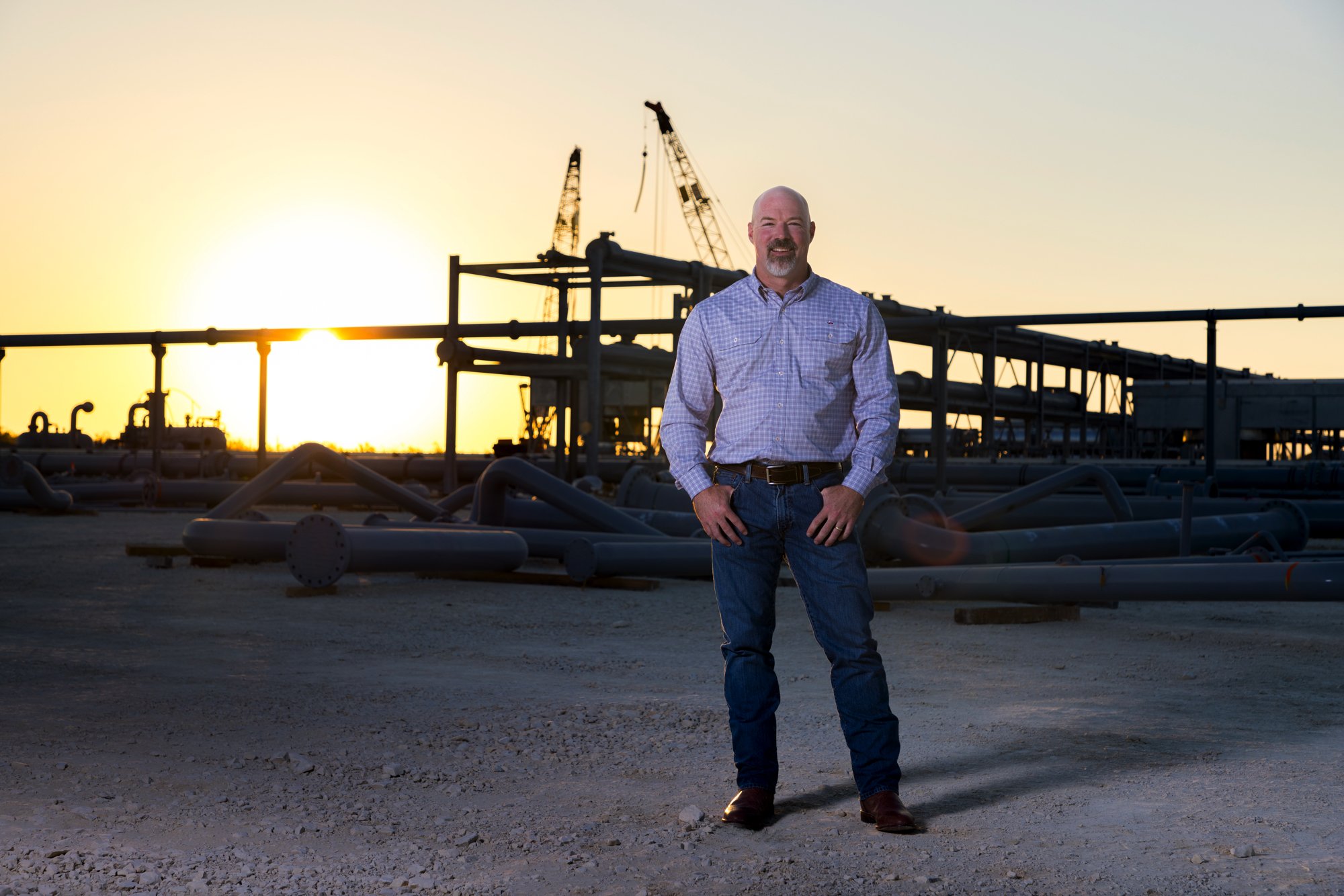 A man standing with hands on hips in front of industrial pipes and equipment at sunset.