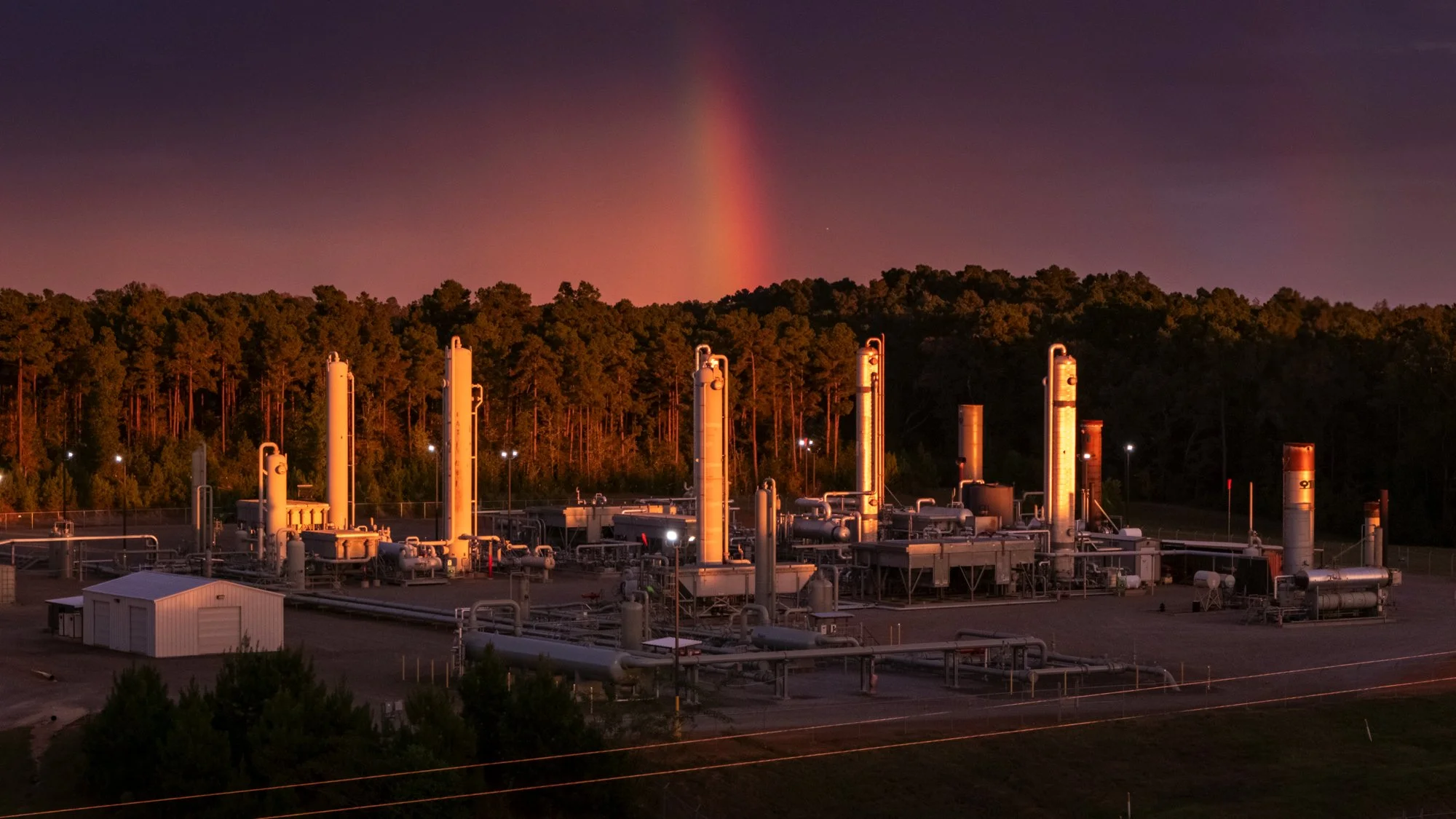 Industrial facility with tall vertical pipes at sunset, with a forest and a rainbow in the background.