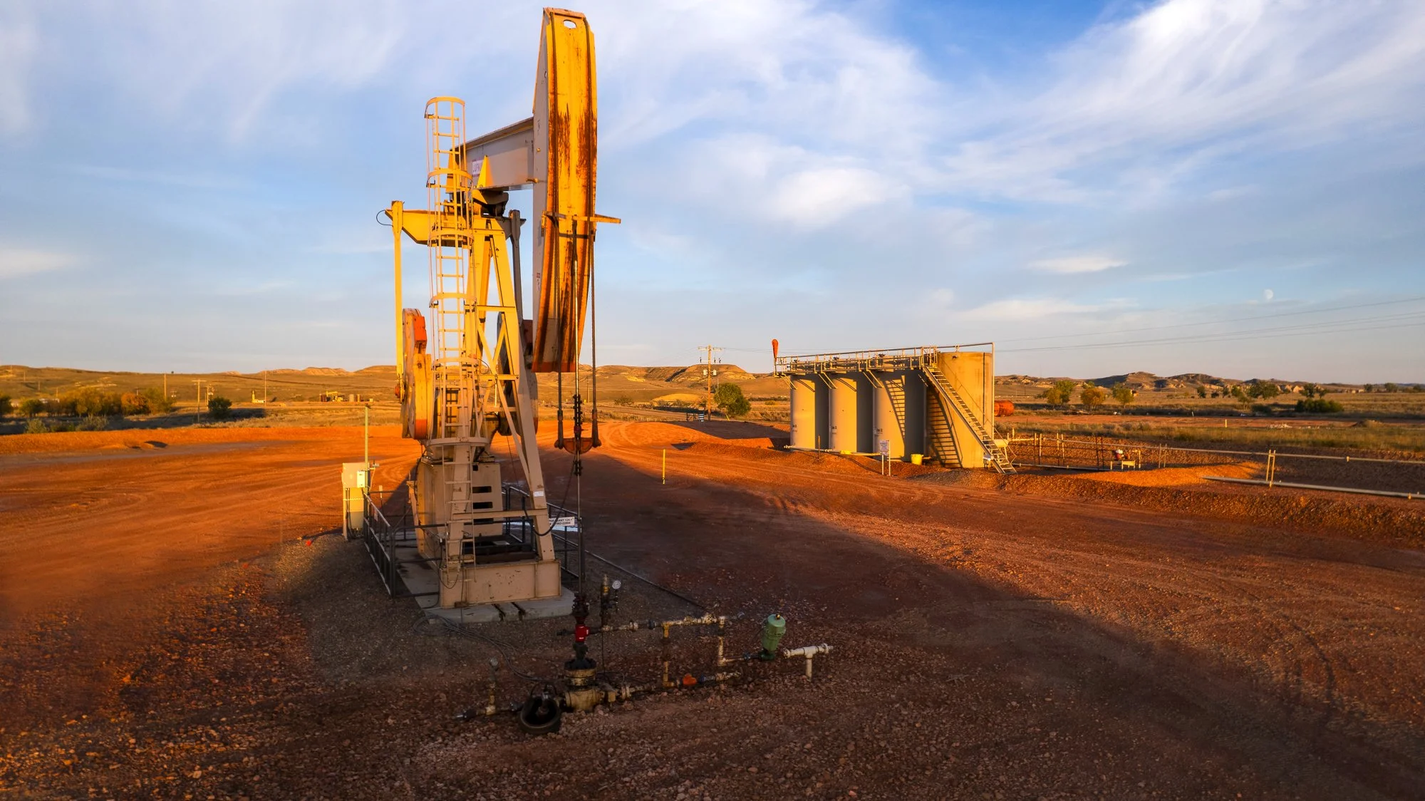 Oil drilling rig on reddish dirt at sunset with pipelines and structures in the background.