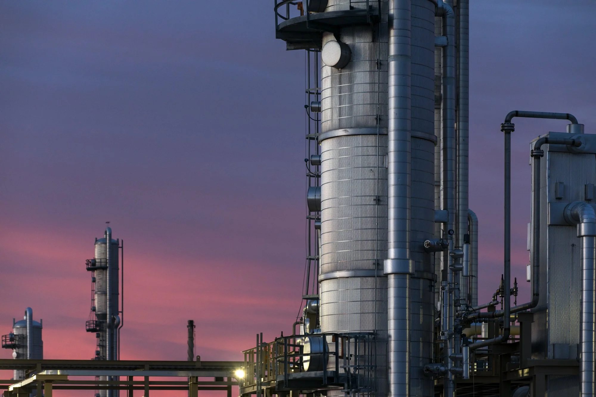 Industrial plant with large silver metal pipes and towers against a pink and purple sunset sky.