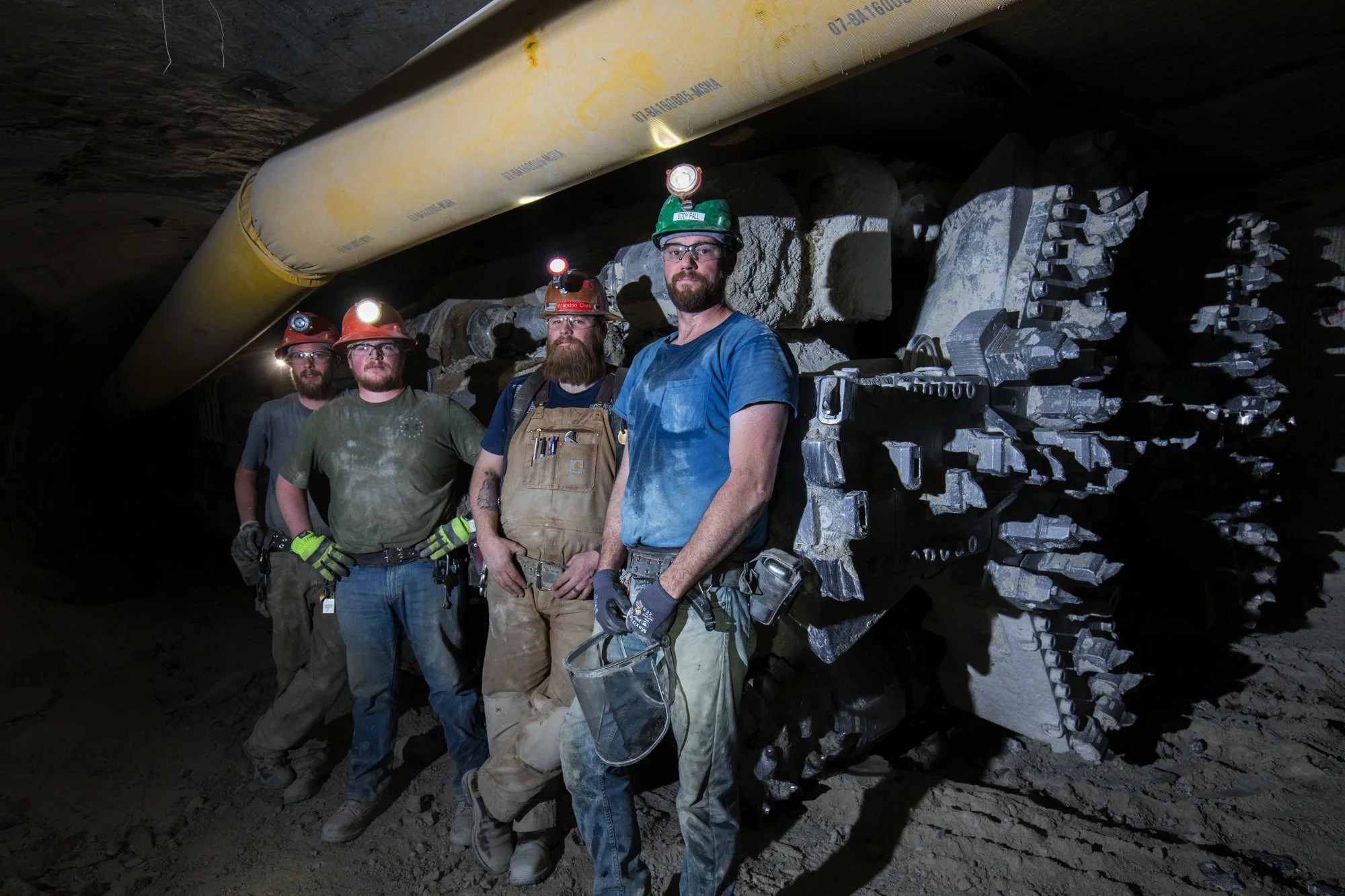 Four miners standing inside a mine tunnel, wearing helmets with lights, work gear, and gloves, with a large mining drill or machine behind them.