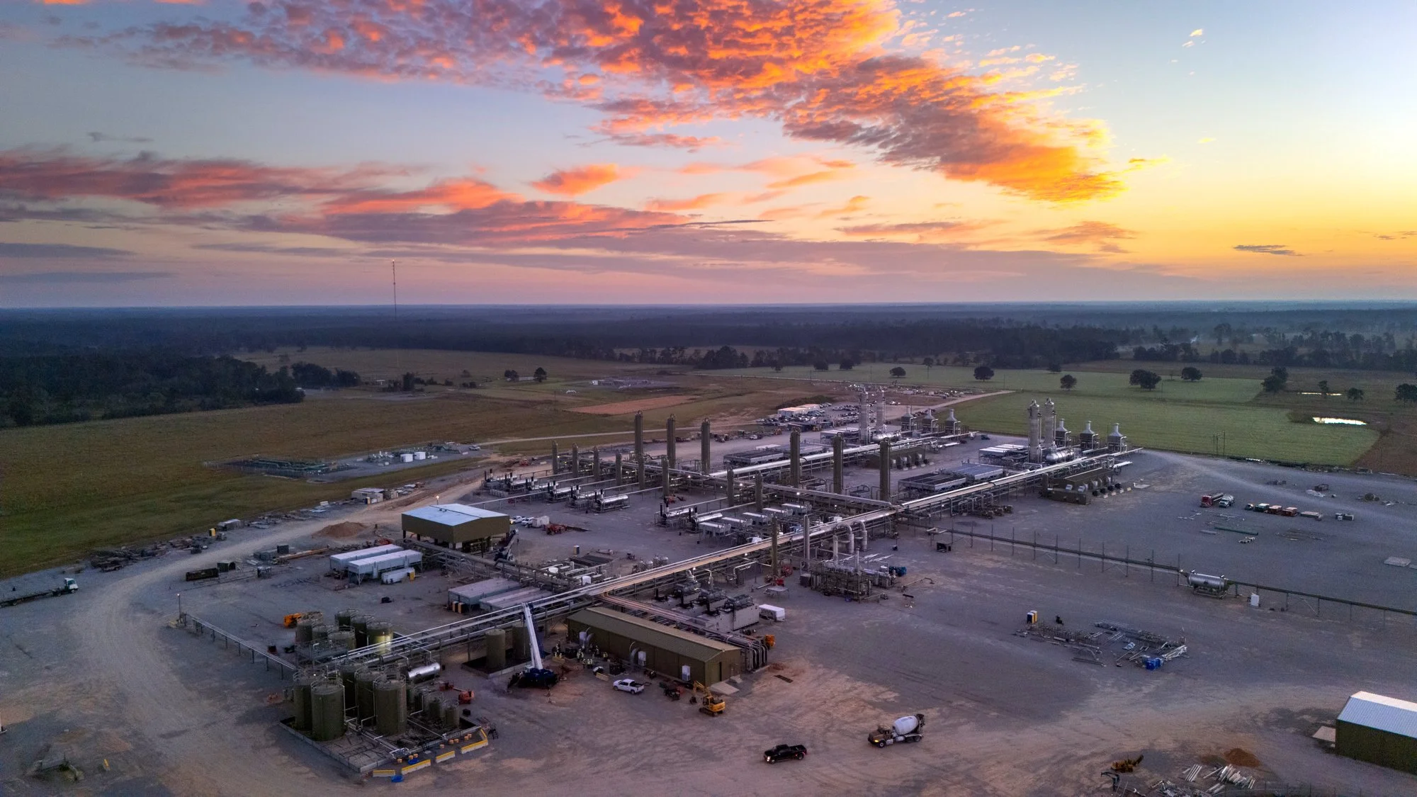 An industrial facility with multiple pipelines and tall structures set in a rural landscape during sunset.