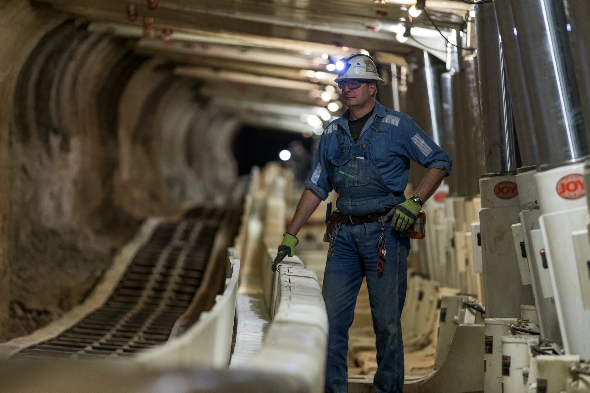A construction worker wearing a safety helmet, glasses, and gloves stands inside a tunnel, inspecting construction equipment along the side.