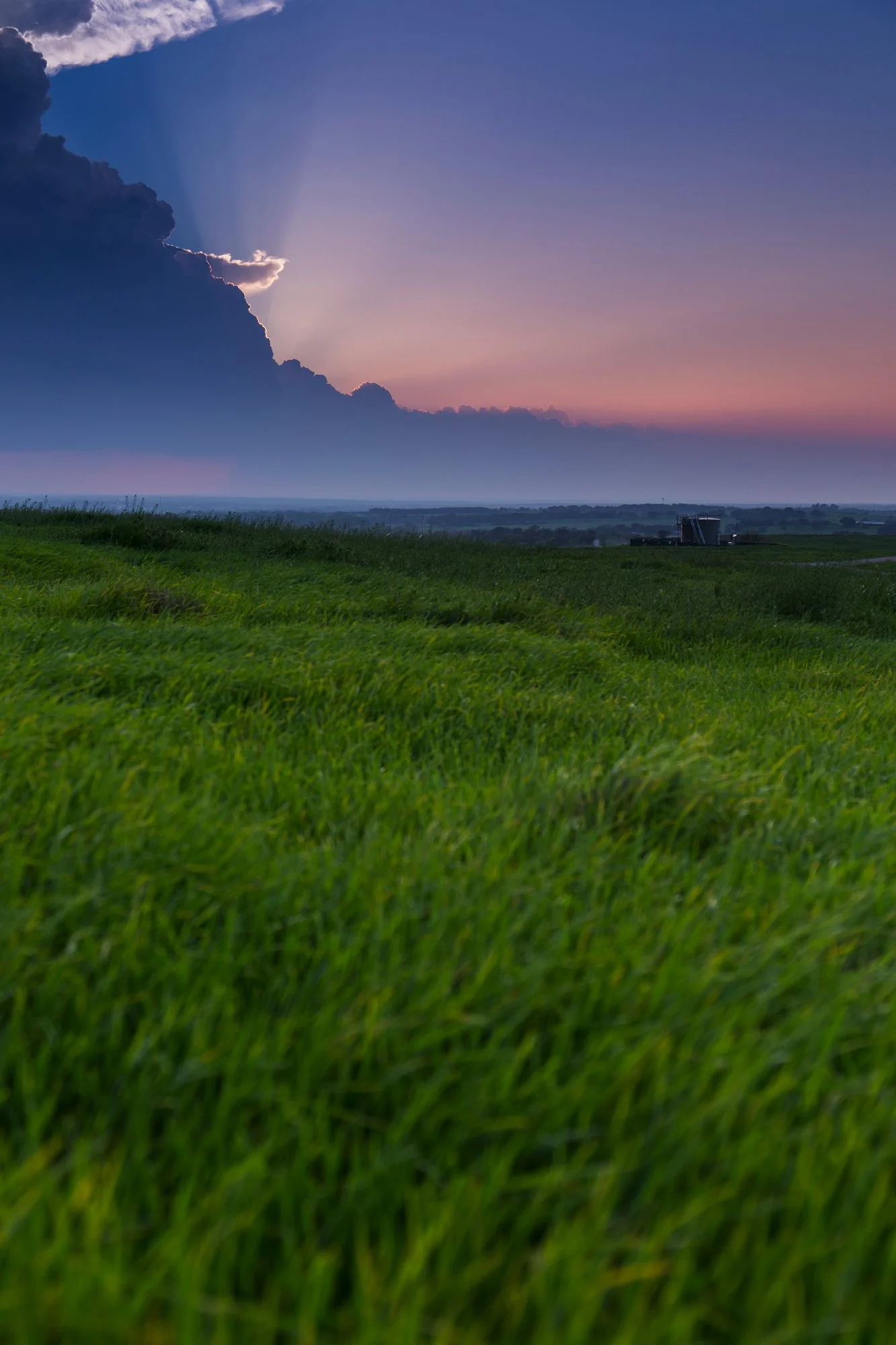 A scenic landscape with lush green grass in the foreground and a distant building on the horizon. The sky is partly cloudy with rays of sunlight breaking through the clouds, creating a soft, colorful sunset or sunrise effect.