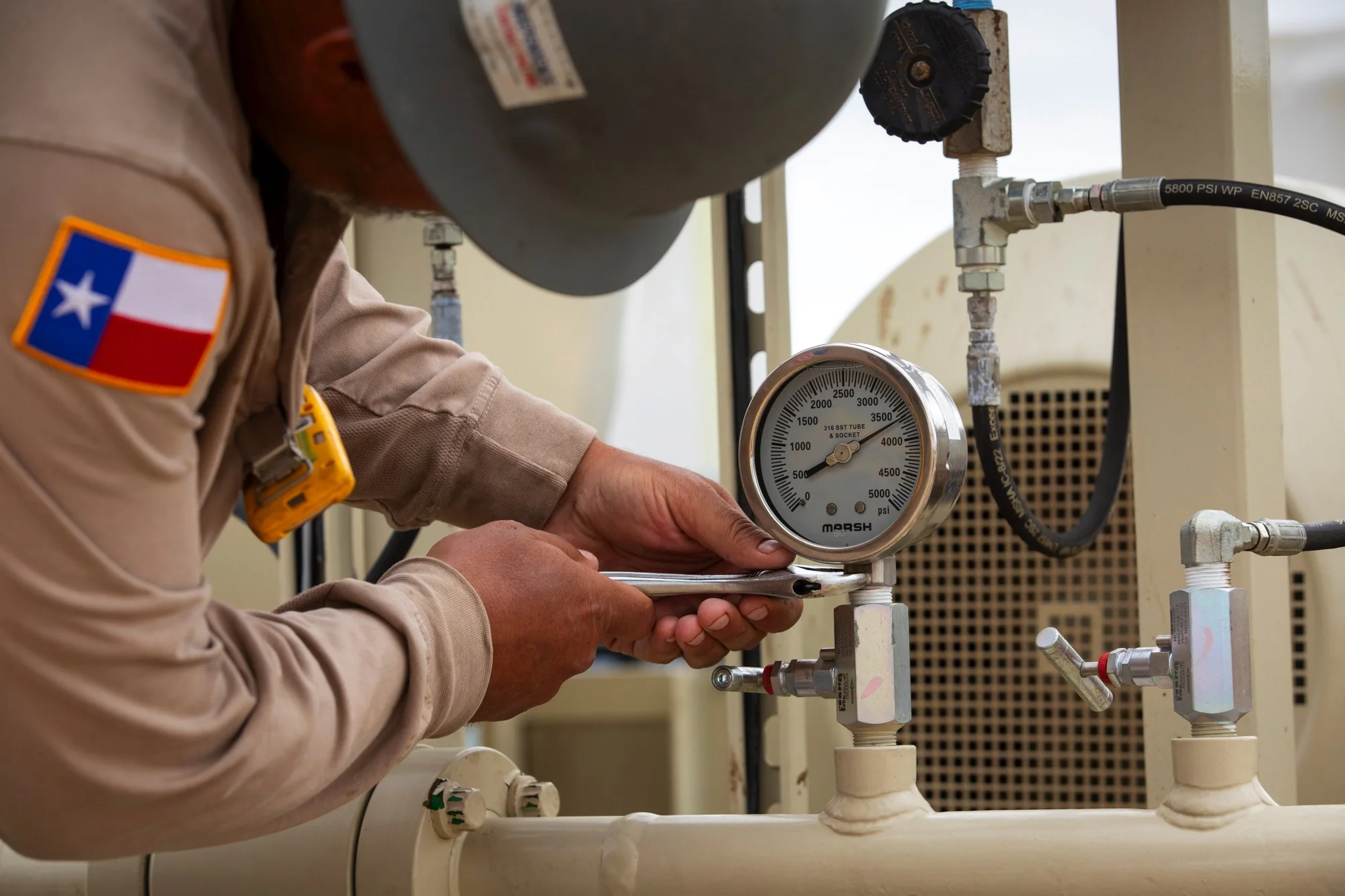 A worker in a uniform with a Texas flag patch is using a wrench to adjust a pressure gauge on industrial equipment.
