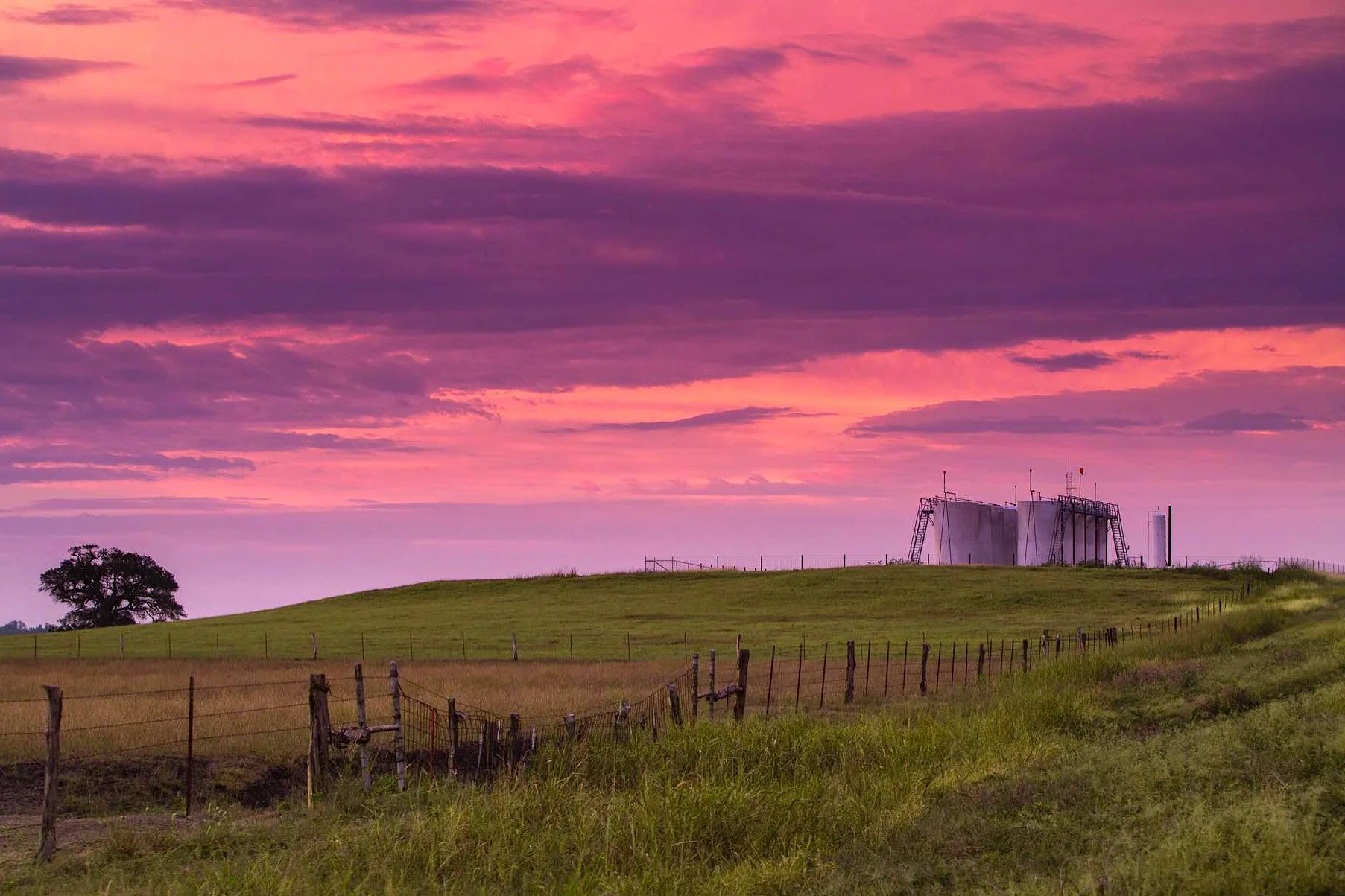A countryside scene with green grassy hills, a wooden fence, a single tree, and industrial tanks under a colorful pink and purple sunset sky.