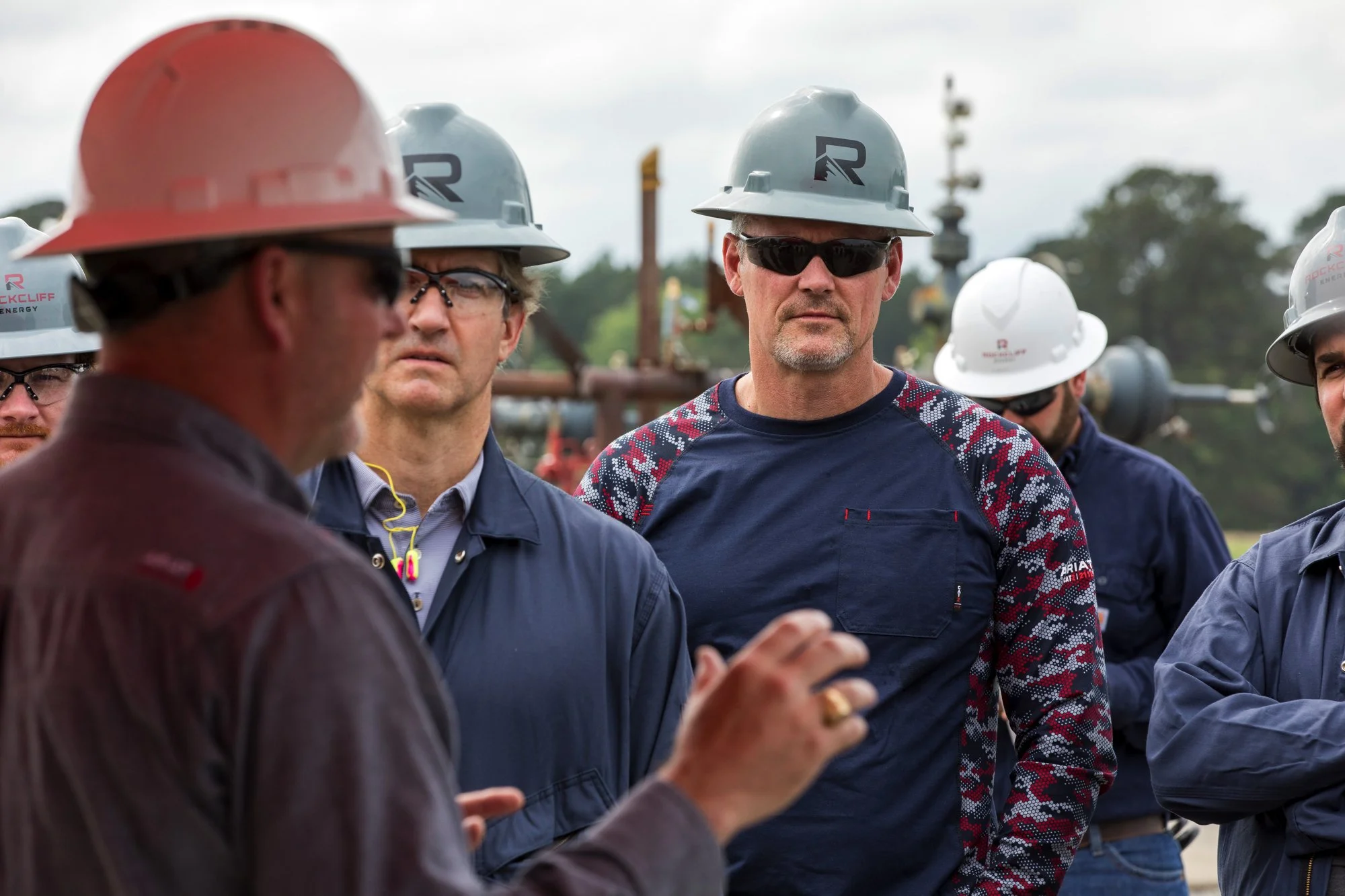 Group of people wearing safety helmets and work jackets, having a conversation outdoors with industrial equipment in the background.