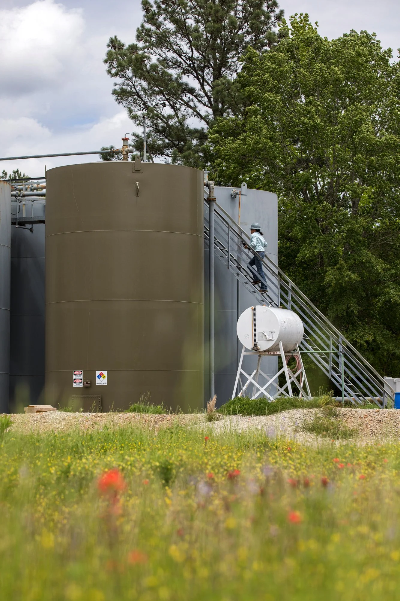 A person in a helmet and light-colored shirt walking down the stairs beside large industrial tanks, with trees and cloudy sky in the background.