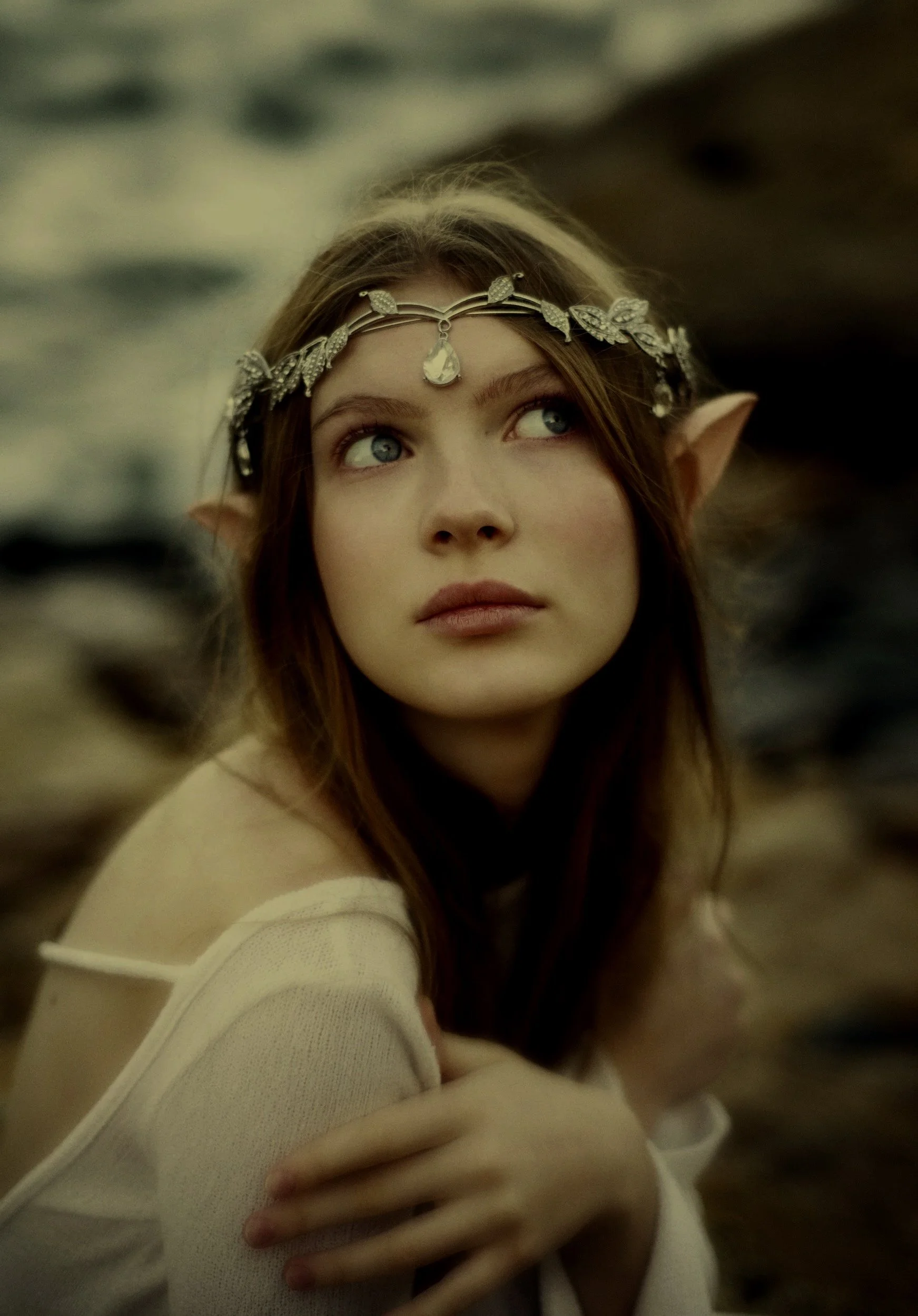 A woman with elf ears and long hair wears a floral headband with a teardrop pendant. She is looking upward, with her hand on her arm, and is dressed in a cream-colored top. The background is blurred with neutral tones.
fantasy ghostwriti
Ghostwriting