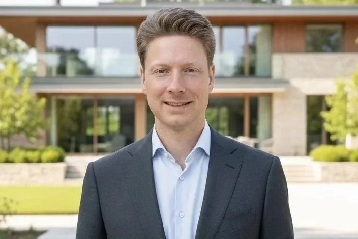 Ein junger Mann in einem dunklen Anzug und hellblauem Hemd steht vor einem modernen Gebäude mit viel Glas und umgeben von Grünflächen.