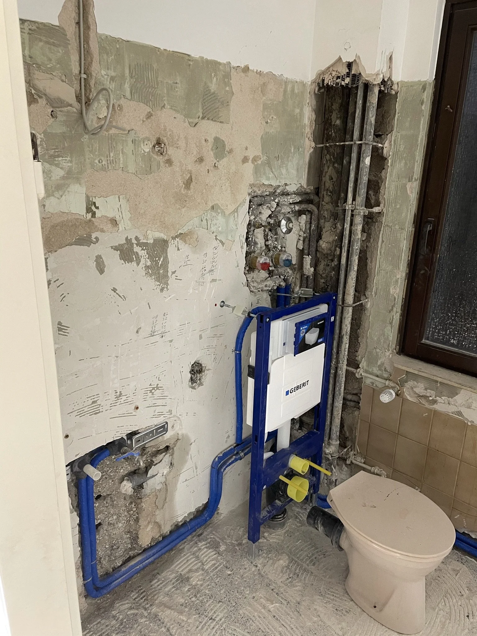 Badezimmer im Umbau mit installiertem Wasser- und Abwasserleitungssystem, vorbereiteter Raum für eine Toilette und eine Wand mit verdeckten Installationen.