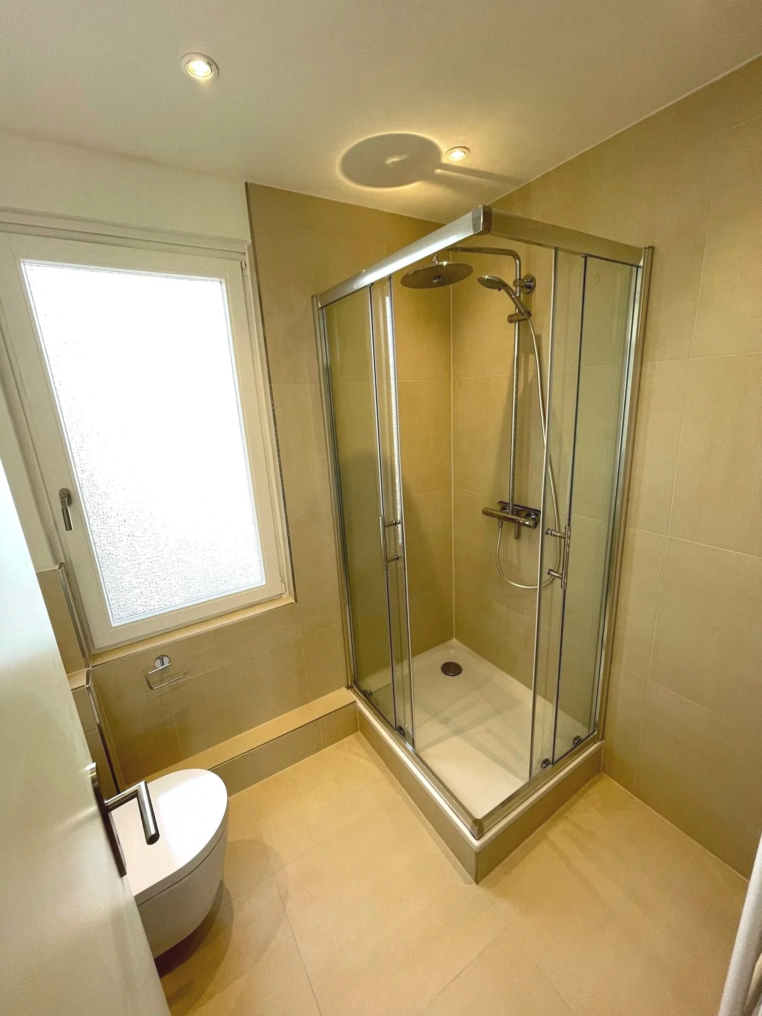 Badezimmer mit Dusche, Fenster und Toilette, beige Fliesen, moderne Ausstattung.