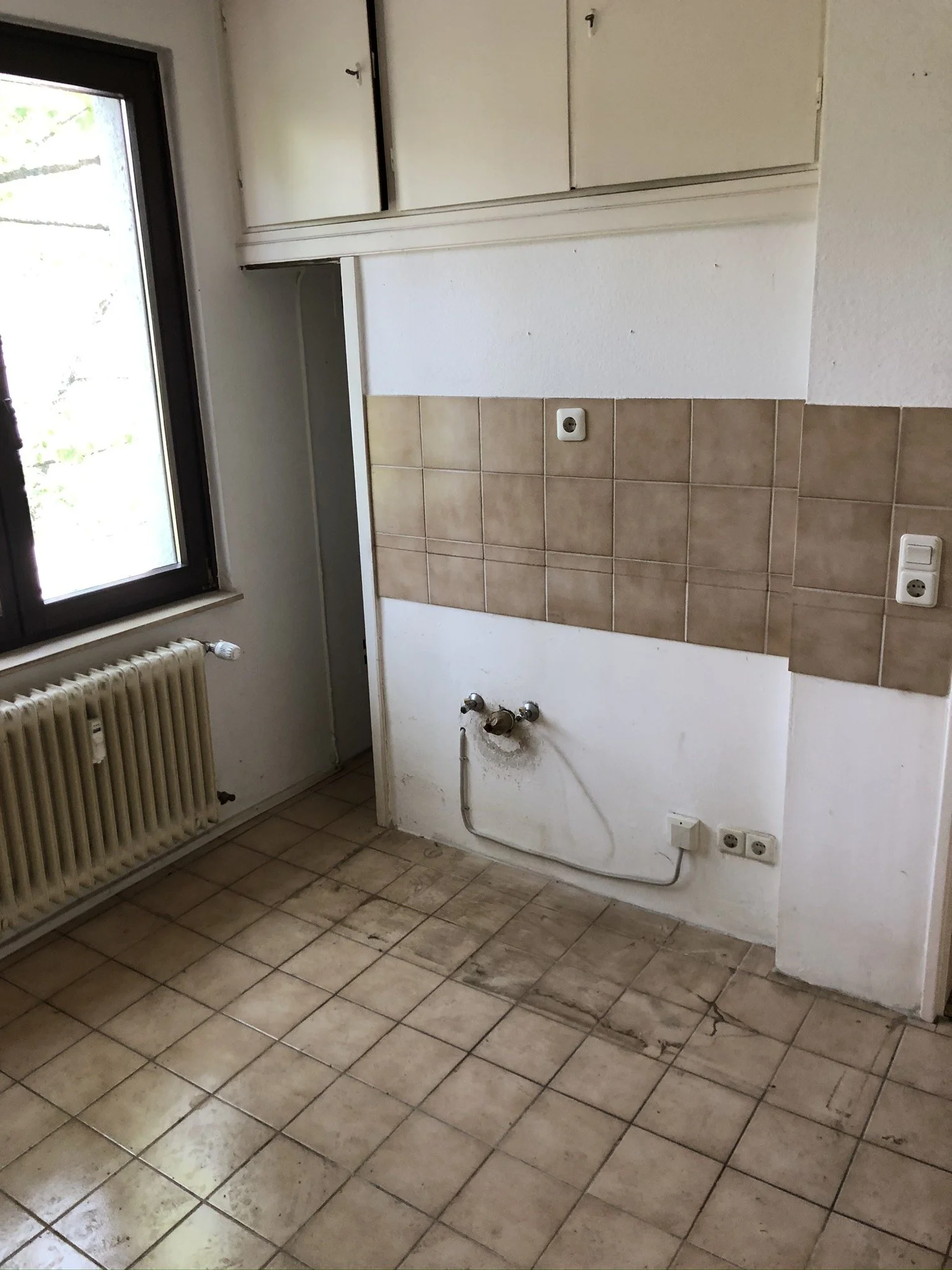 Ein leerer Küchenbereich mit Fliesenboden, einer Wand mit braunen Fliesen im oberen Bereich, einer Wasseranschlussleitung für eine Spüle, einer elektrischen Steckdose und einem Fenster auf der linken Seite. Oben befinden sich weiße Küchenoberschränke.