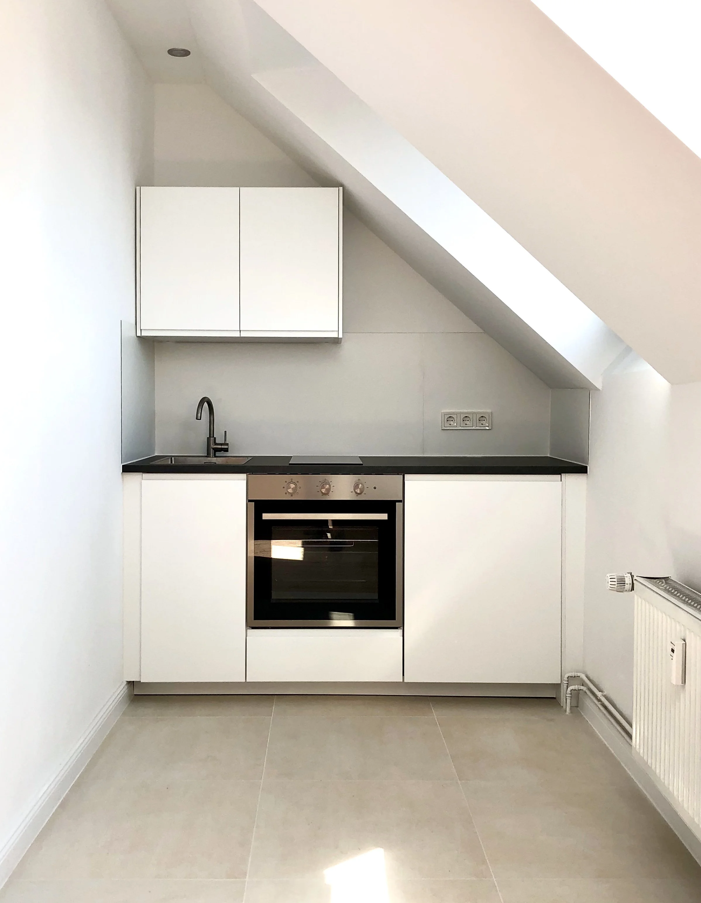 Küche mit weißen Schränken, schwarzen Arbeitsfläche, kleiner Spüle, Backofen, unter Dachschräge, helle Wandfarben und Fliesenboden.