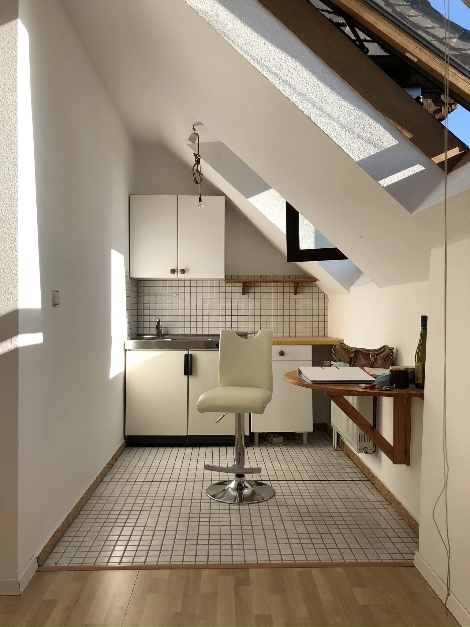 Ein kleines Küchenstudio im Dachgeschoss mit weißen Schränken, einer kleinen Küchenzeile, einem weißen Barhocker und einem Tisch mit Akten, einer Flasche Wein und anderen Gegenständen. Es gibt ein Dachfenster und eine Holzdecke.