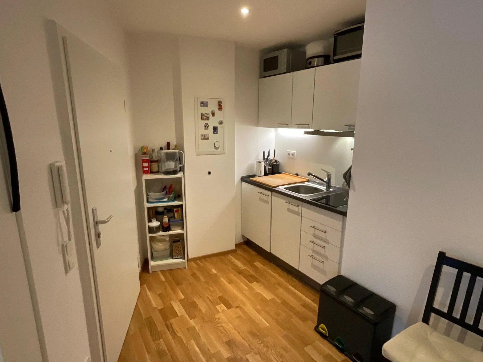 Kleine Küche mit weißen Schränken, Spüle und Herd, Holzfußboden, kleine Regale mit Lebensmitteln und Küchenutensilien, schwarzes Mülltrennsystem, Sitzgelegenheit mit Stuhl, beleuchtete Arbeitsfläche.