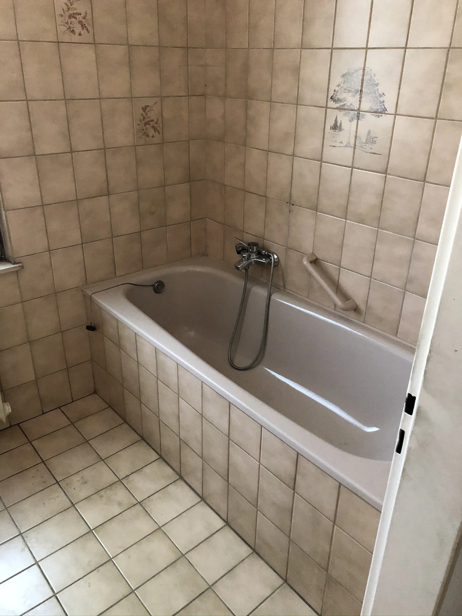 Ein Bad mit beige Kacheln, eine Badewanne mit einem Wasserhahn und Duschkopf, eine seitliche Haltestange an der Wand, einige beschädigte Kachelbilder an der Wand.
