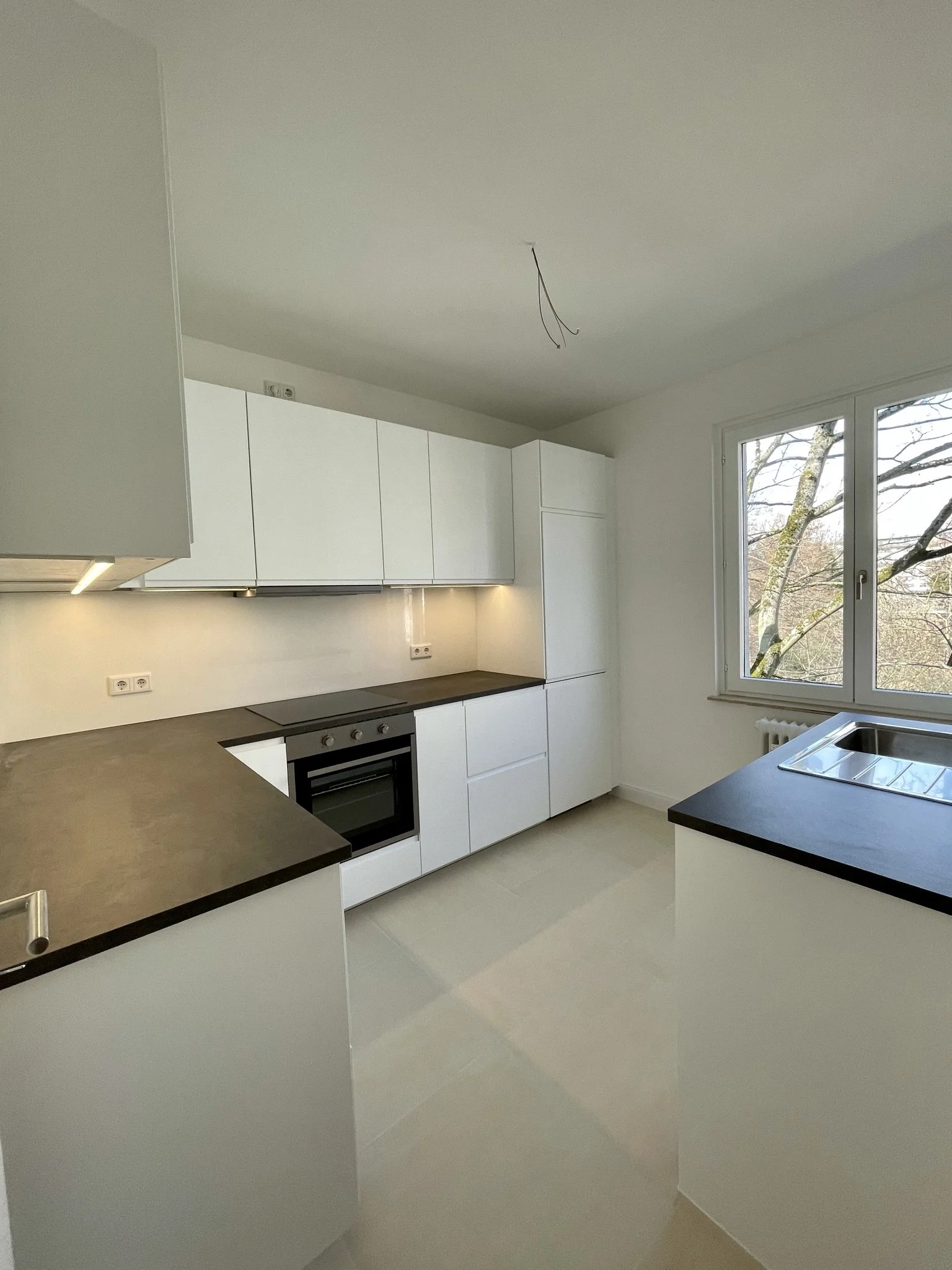 Moderne weiße Küche mit schwarzen Arbeitsplatten, Herd, Spüle und großem Fenster mit Blick auf Bäume.