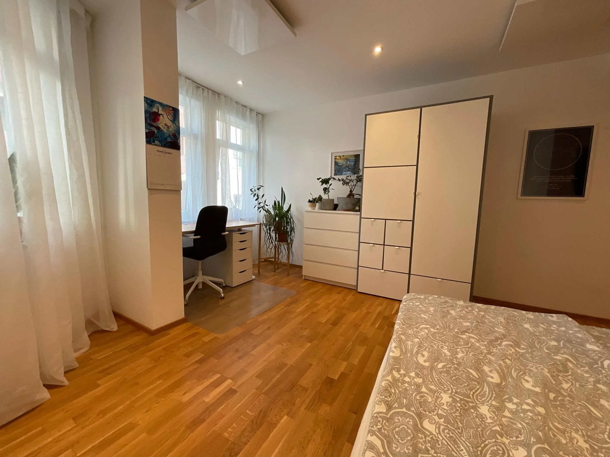 Ein Schlafzimmer mit Holzboden, weißen Vorhängen und einem Bett. Im Hintergrund ist ein Schreibtisch mit einem schwarzen Stuhl, Pflanzen und einem weißen Schrank. An der Wand hängen Bilder.