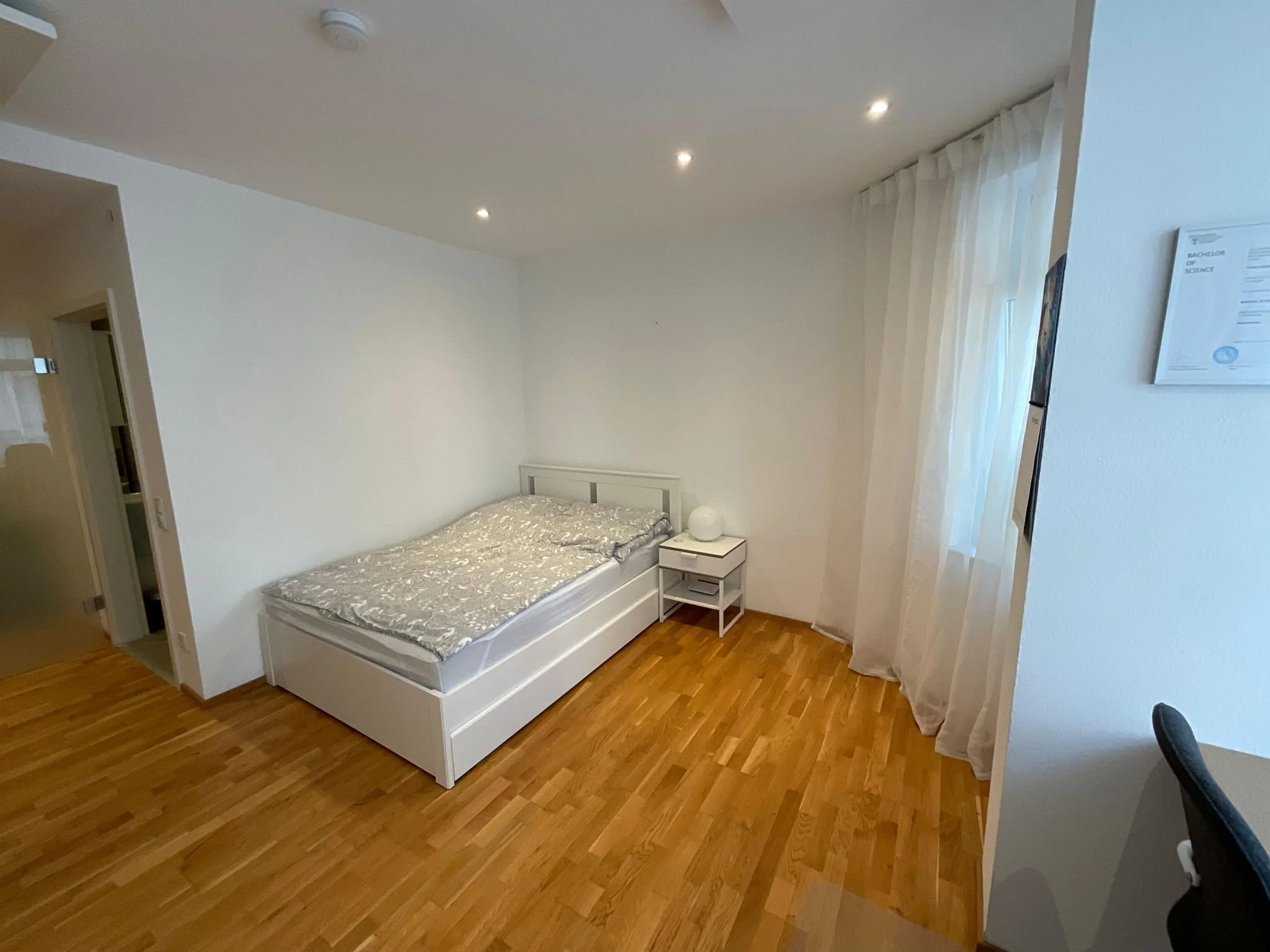 Ein schlichtes Schlafzimmer mit weißen Wänden, Holzboden, einem weißen Bett, Nachttisch mit Lampe und einem Fenster mit weißen Vorhängen.