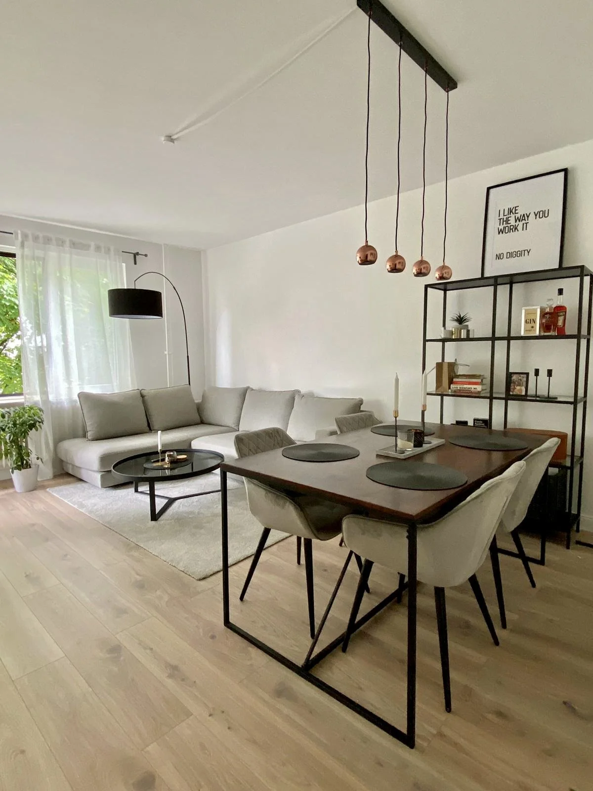 Modernes Wohnzimmer und Essbereich mit weißem Sofa, dunklen Couchtisch, Esstisch mit sechs Stühlen, schwarzen Regal mit Dekorartikeln, Wandbild, Lampe mit schwarzen Lampenschirm, große Fenster mit weißen Vorhängen, Holzboden und Pflanzen.