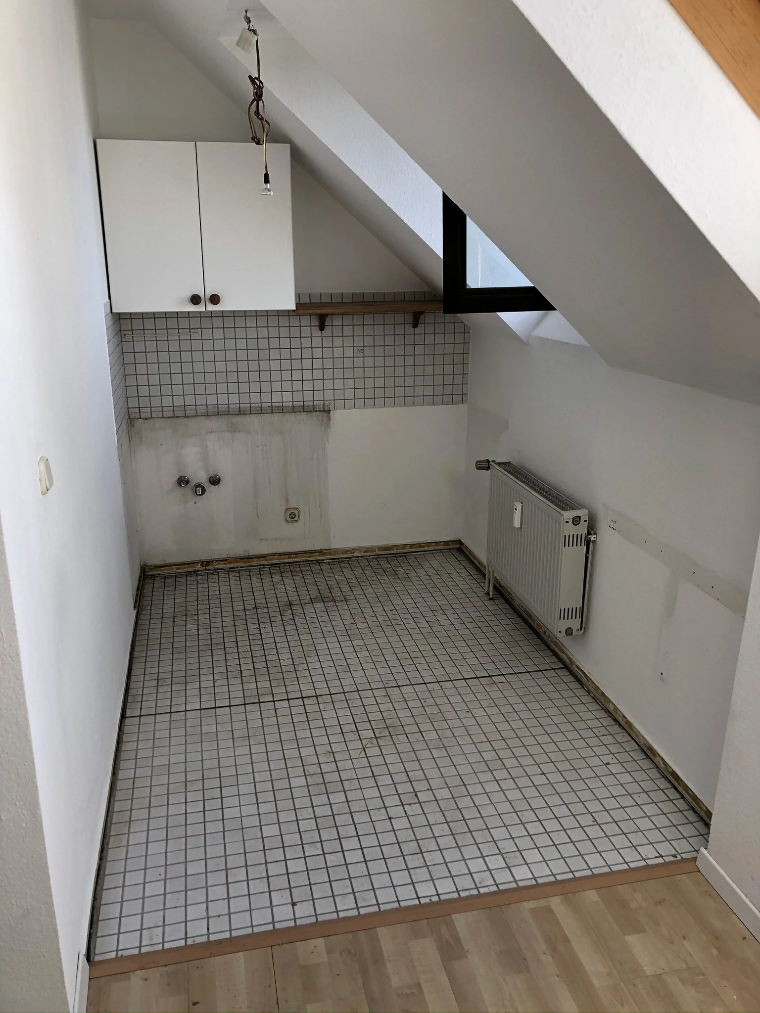 Unvollständige Küche mit weißen Fliesen, einem Heizkörper, einer weißen Hängeschrank und einem Fenster unter dem Dach, noch ohne Küchenmöbel und Geräte.