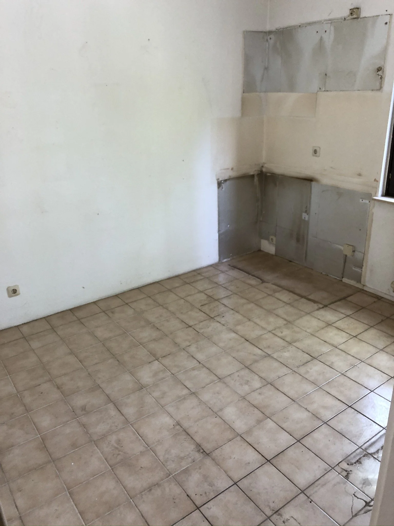 Ein leerer, unrenovierter Raum mit beige Fliesenboden und weißen Wänden, eine Wand ist teilweise mit Metallplatten bedeckt.