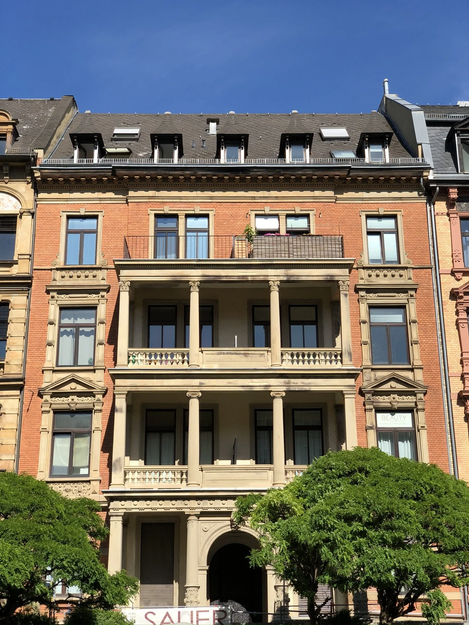 Mehrstöckiges Gebäude mit roten Backsteinwänden, klassischen Säulen auf den Balkonen und grünen Bäumen im Vordergrund