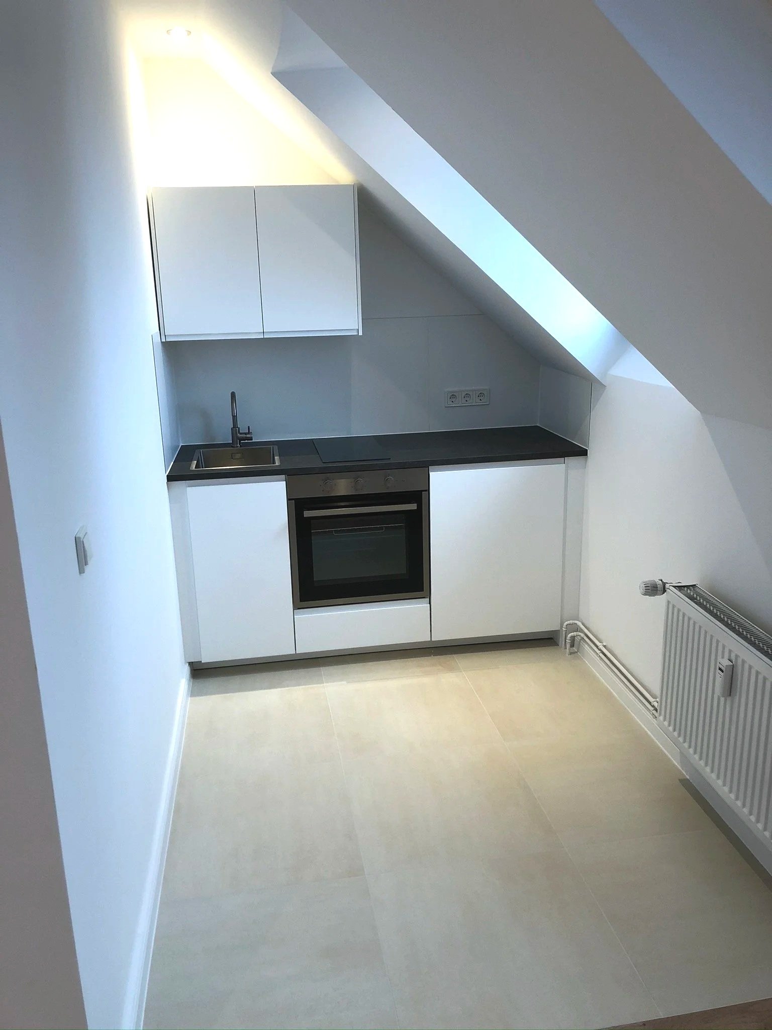 Kleine moderne Küchenzeile mit weißen Schränken, dunklem Arbeitsplatten, Backofen, Spüle, unter einem Dachfenster