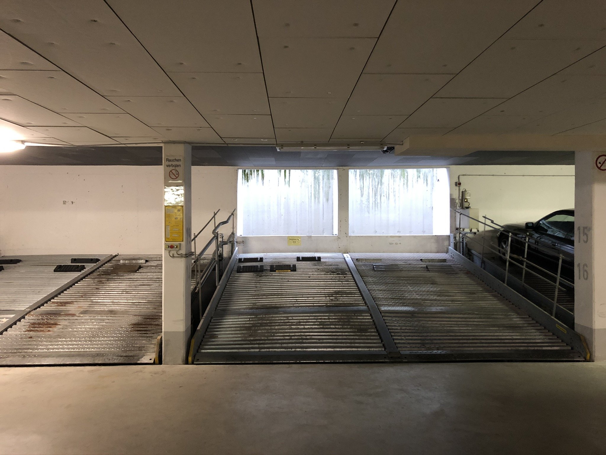 Leerer Parkausgang in einer Tiefgarage mit einem rollbaren Parkplatzsystem, eine Wand im Hintergrund mit einem Wasserfall und einem vorgeparkten schwarzen Auto auf der rechten Seite.