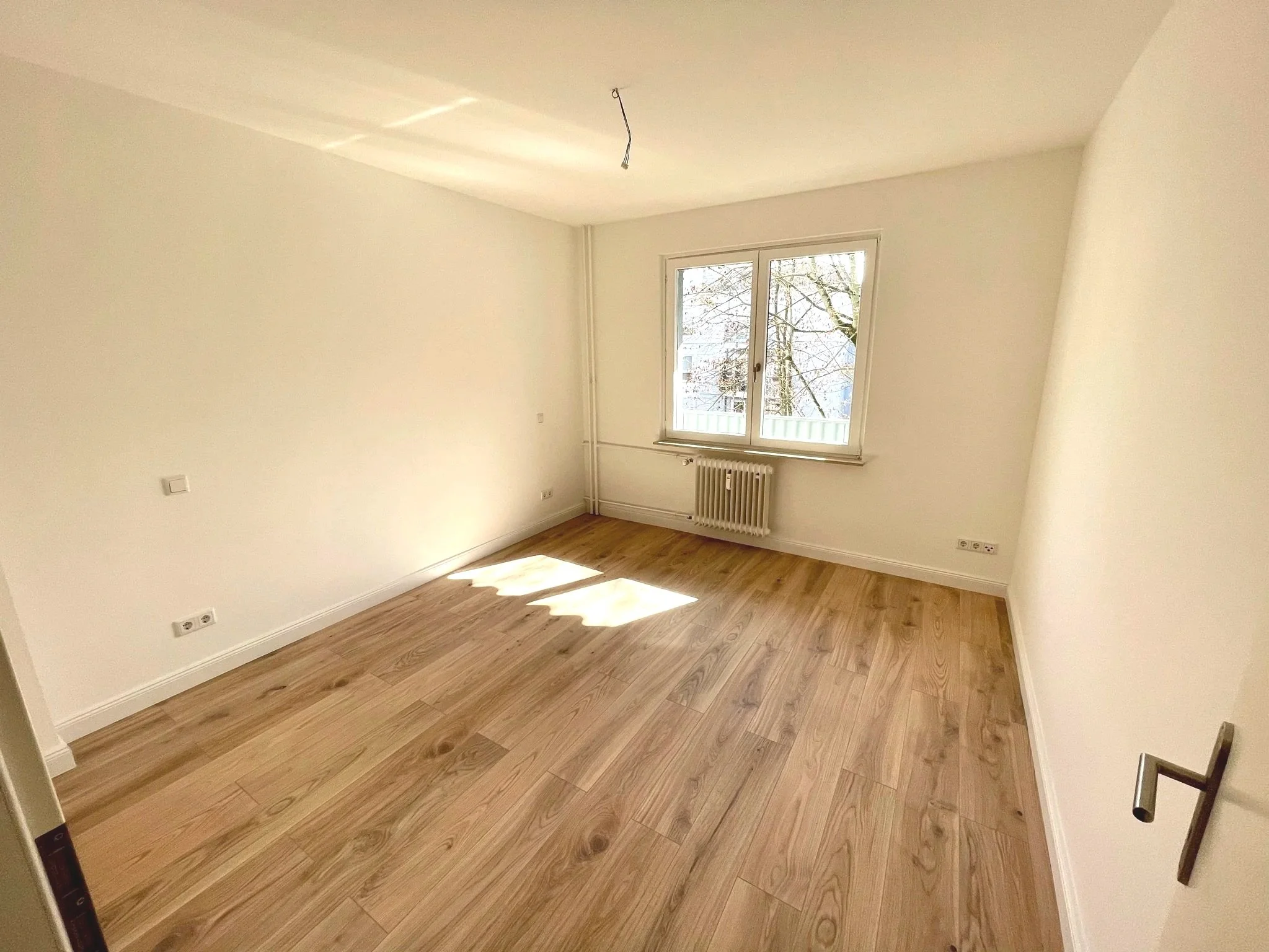 Leeres Zimmer mit Holzfußboden, weiß gestrichenen Wänden, einer großen Fensterscheibe und Heizkörper darunter.