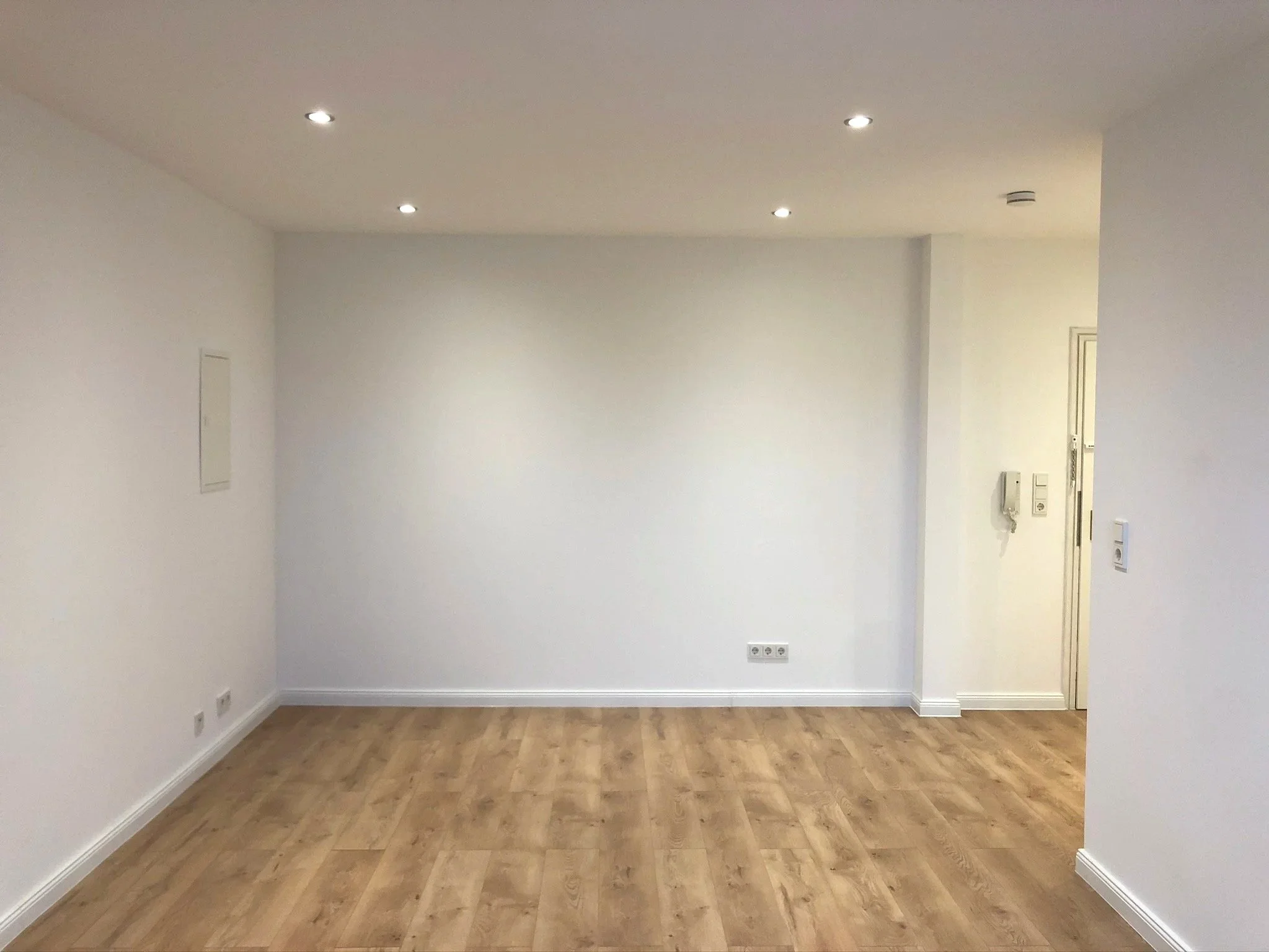 Leeres Wohnzimmer mit weißen Wänden, Holzboden, Deckenlichtern und einem Flur auf der rechten Seite.
