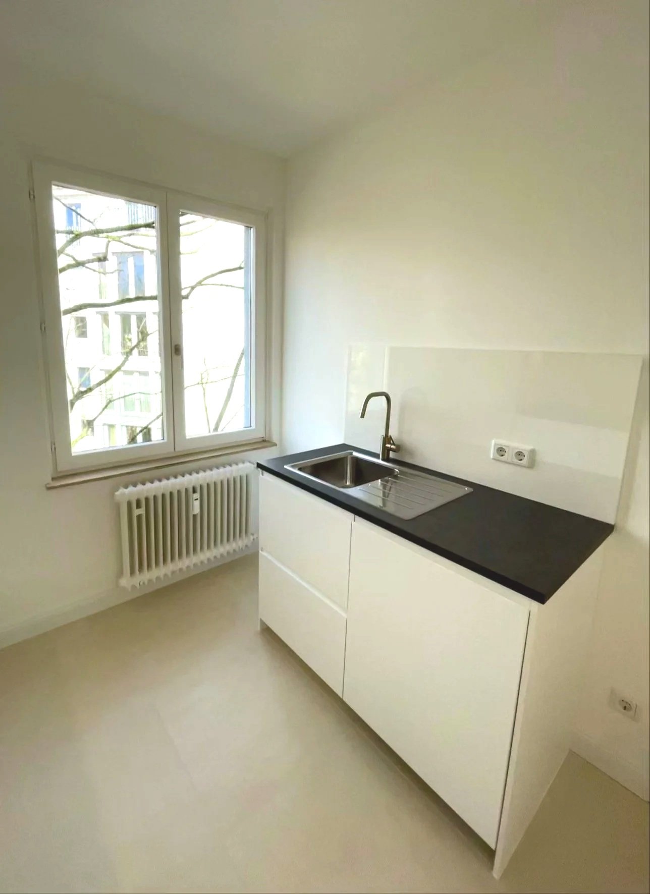 Kleine Küche mit weißem Unterschrank, schwarzer Arbeitsfläche, Edelstahlspüle und Wasserhahn, Fenster mit Blick auf Bäume, Heizkörper unter dem Fenster, weiße Wände und Boden.