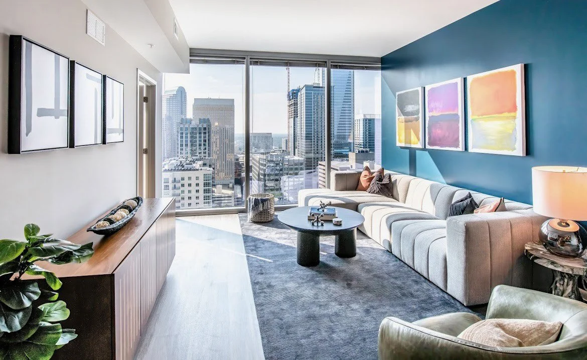 Living room with large windows showing city skyscrapers, modern white sofa with various pillows, round black coffee table, abstract colorful paintings on a blue accent wall, side table with lamp, and a console with decorative items.