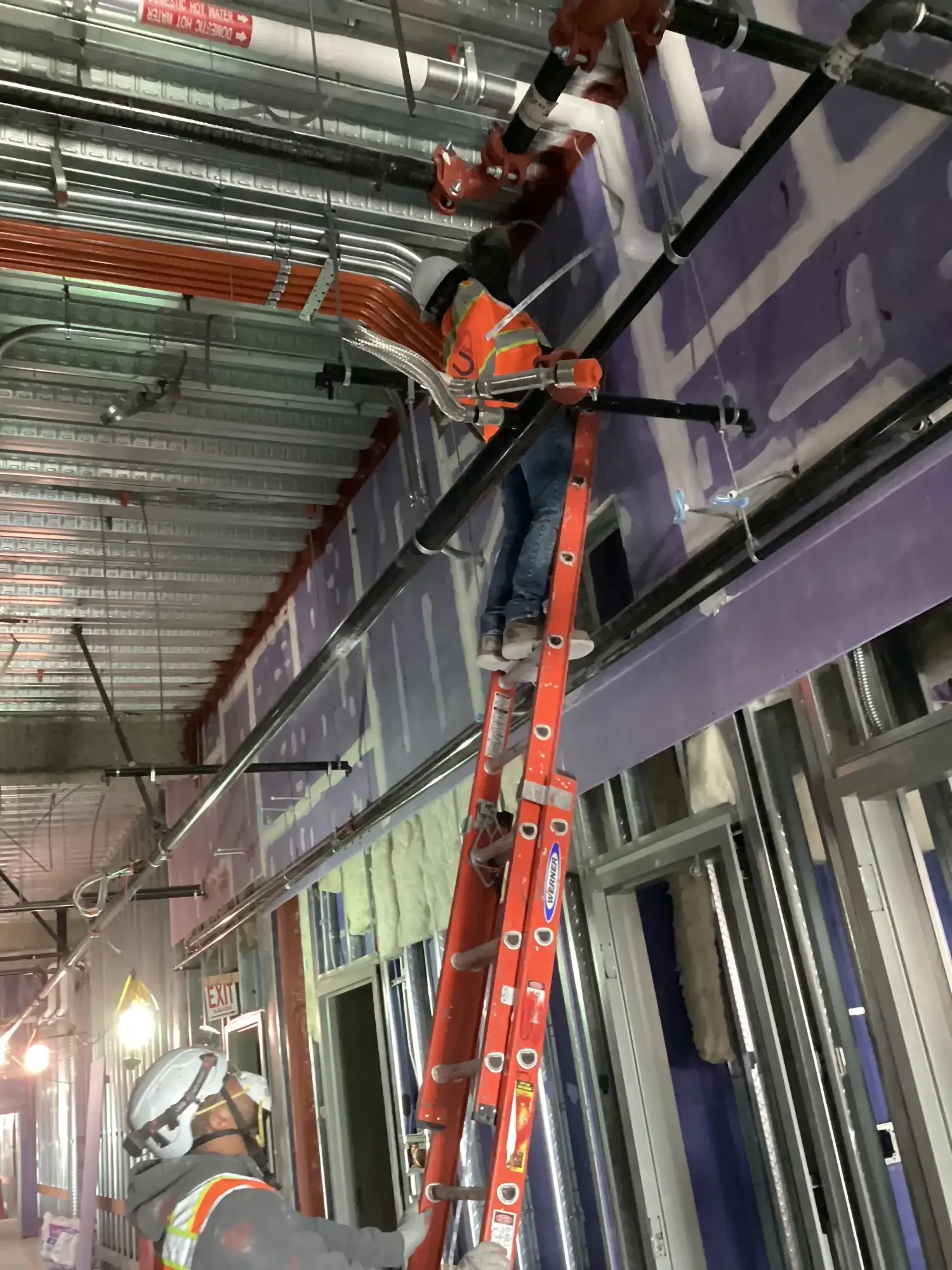 Construction worker on a ladder installing or repairing pipes and wiring inside a building under construction.